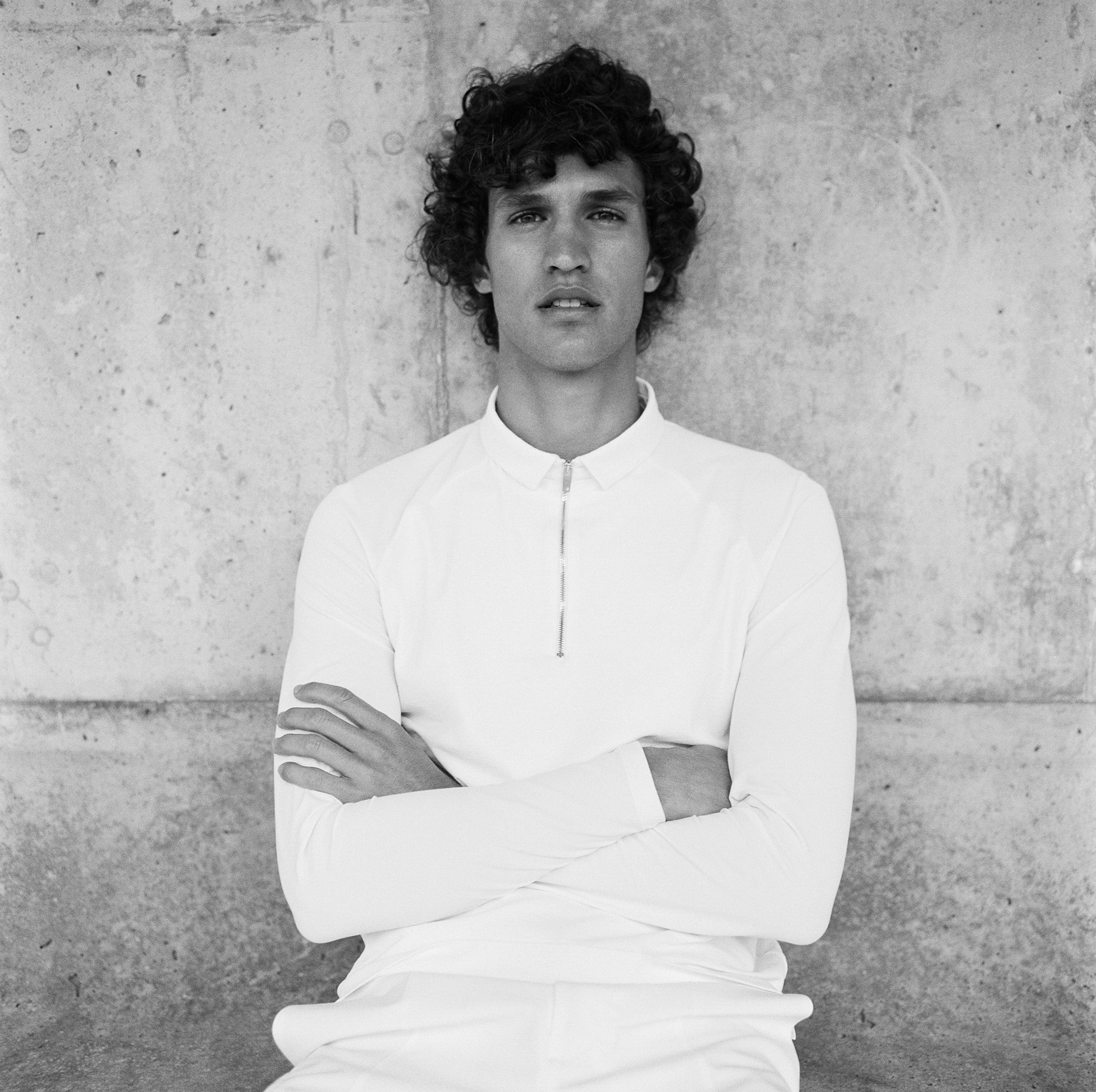 A man with curly dark hair is leaning against a grey stone wall. He is wearing a long sleeve white shirt with a zipper. He is crossing his arms. 