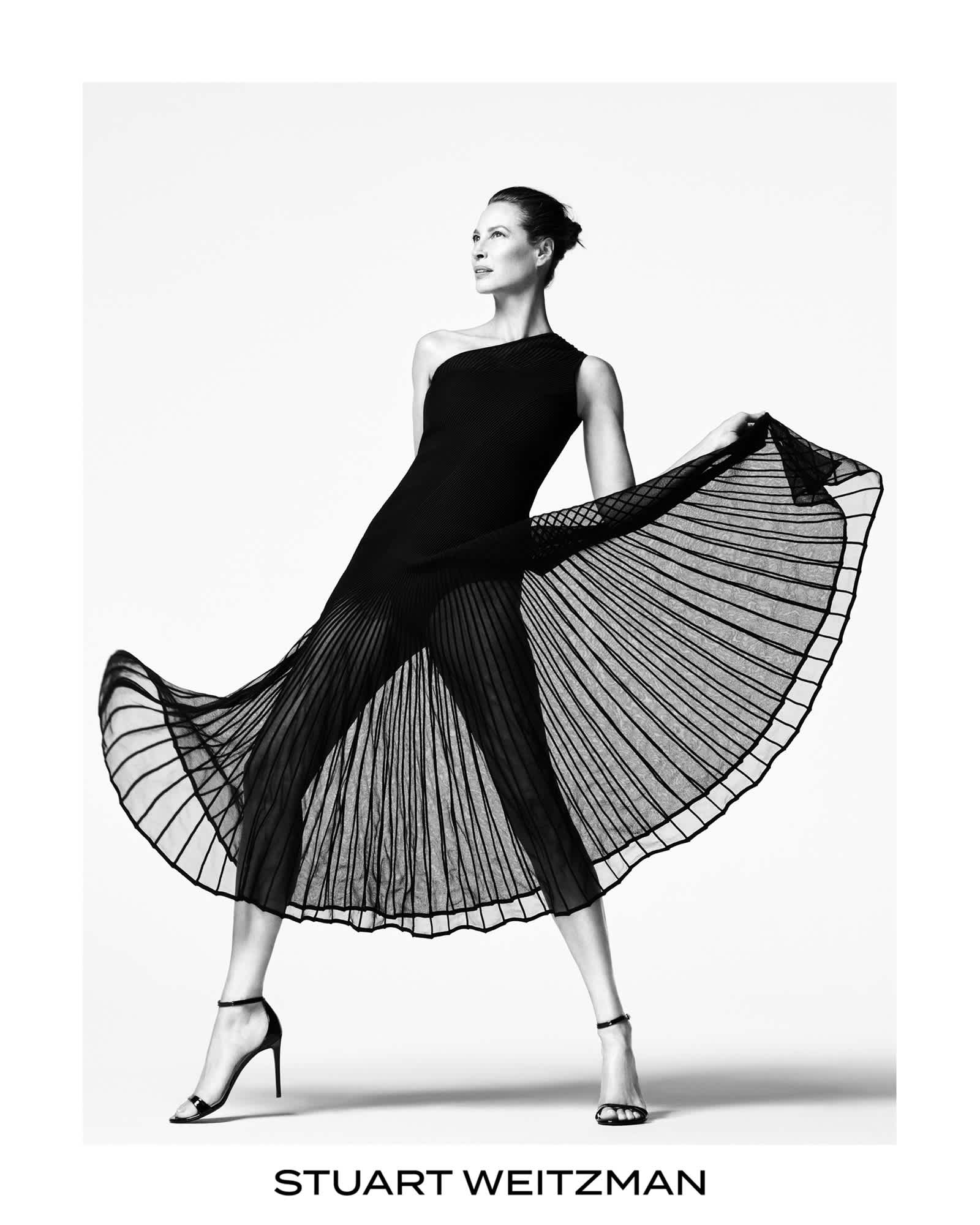 Christy Turlington is lunging forward wearing a semi translucent pleated black dress. She is holding the sides of the dress up, giving the dress more shape and volume. Stuart Weitzman logo is featured on the bottom center of the frame.