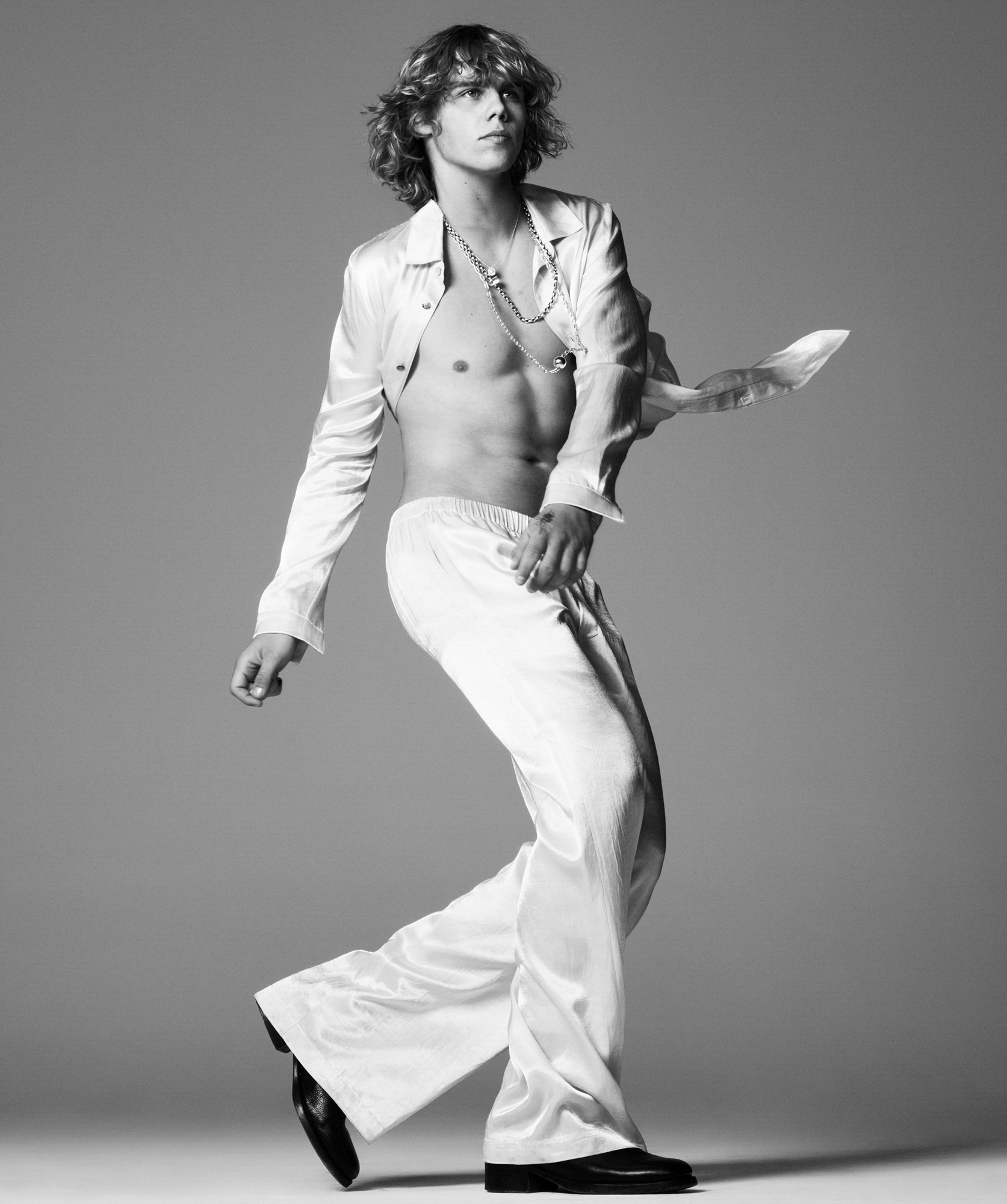 Kid Laroi is wearing an all white look in the center of the frame. He has an ope button up shirt, trousers, and boots. He is in motion stepping forward, has silver jewelry on, and is looking off into the distance past the right side of the frame. 