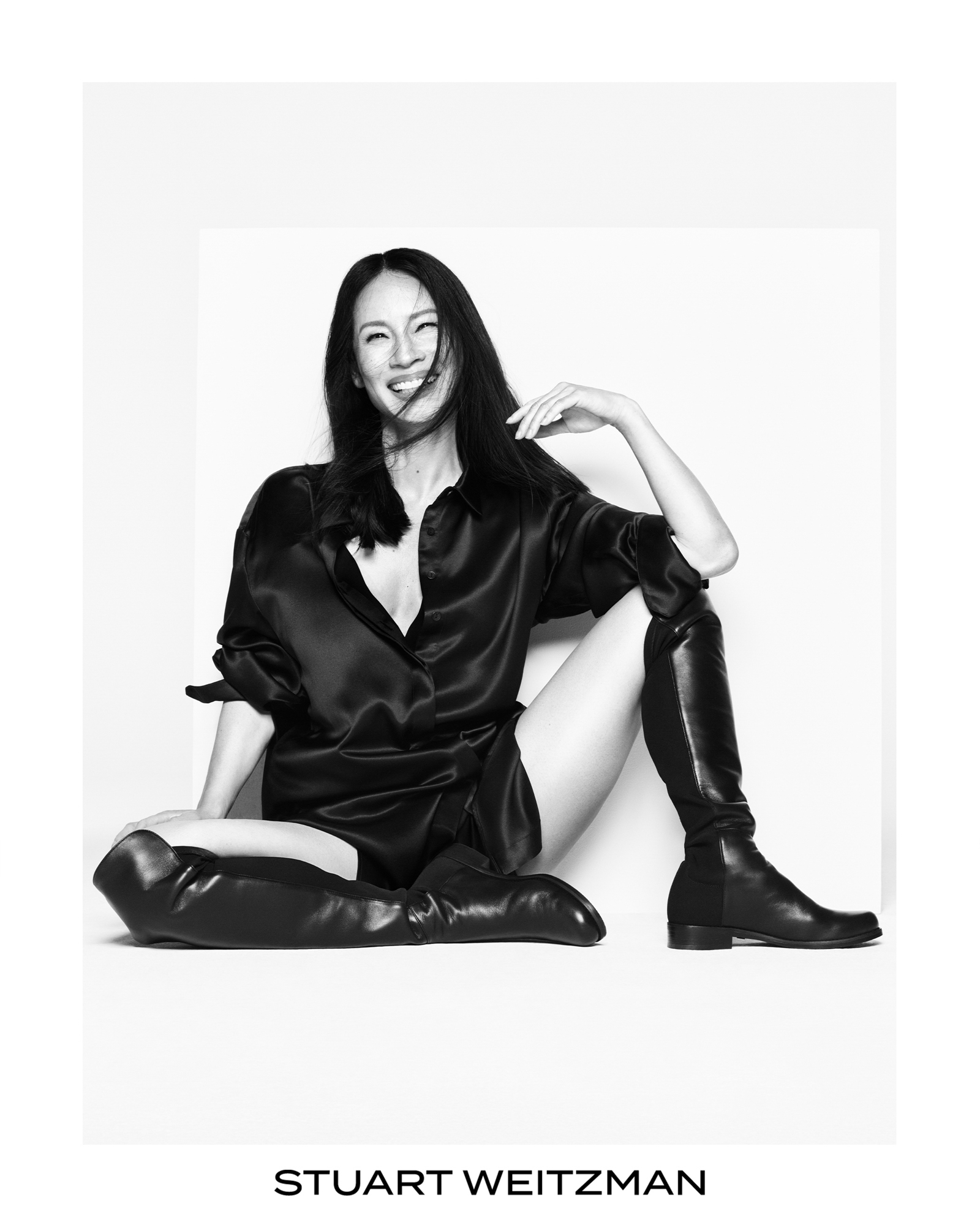 Lucy Liu is seated on the group wearing an all black silky outfit. She is wearing knee length black boots. Lucy is smiling at the camera, and has one of her elbows resting on her knee. Stuart Weitzman logo is featured on the bottom center of the frame. 