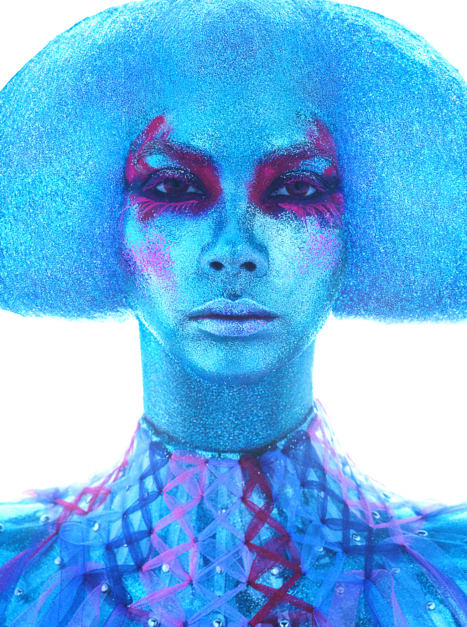 A blue figure fills the frame of the image. The model has a sculpted mushroom look hairstyle, the hair blends with the skin tone which is a blue glittery complexion. The model has a reddish pink eyeshadow look that frames the bottom lash line and extends up above the eyebrows. The neck of the collar has twists going down the neck in shades of reds and pinks. The background of the image is a blown out white. 