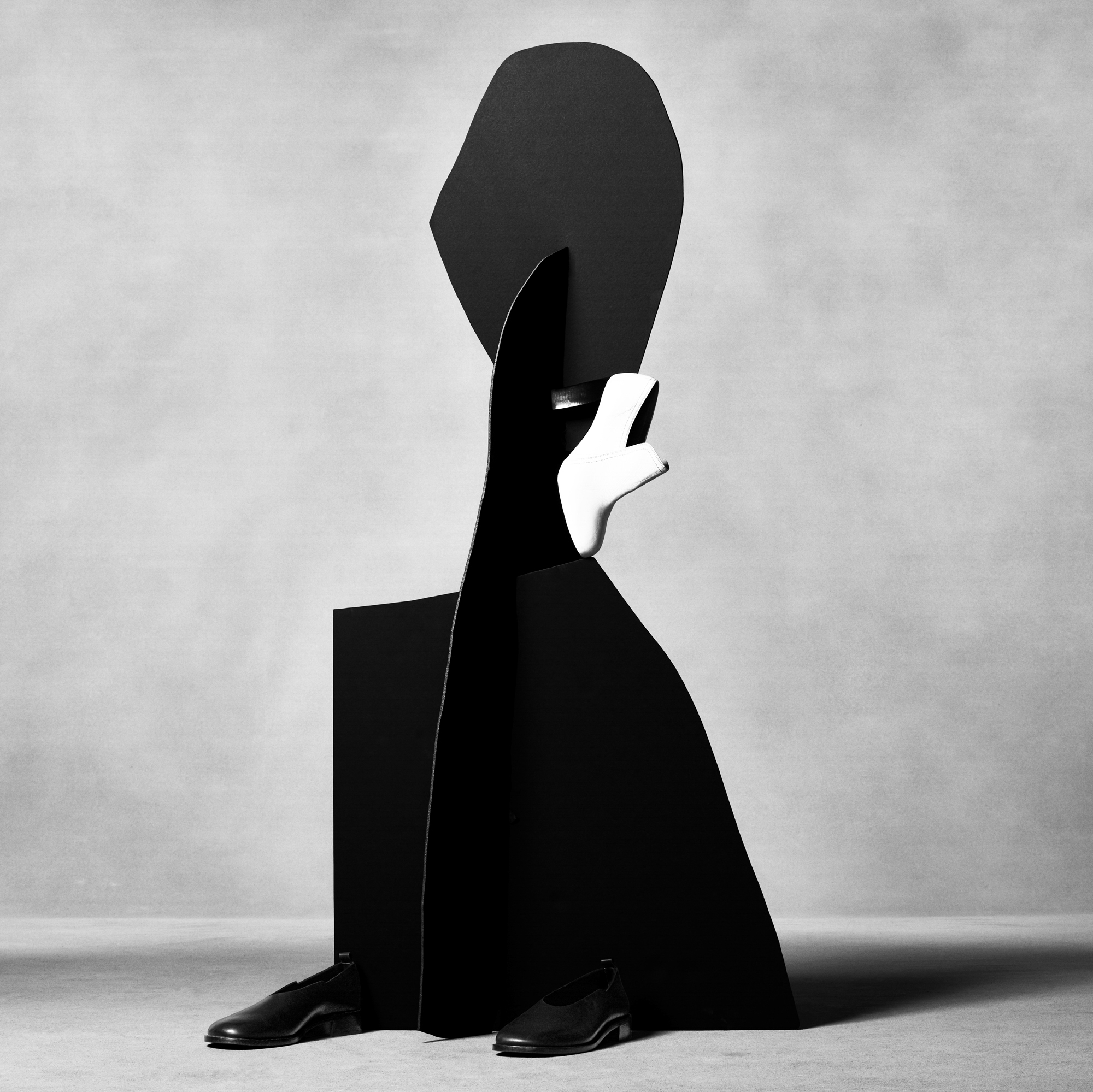 Abstract organic black shapes are connecting in the center of the frame to form a sort of sculpture. Two black shoes are at the base of the sculpture, and a white heel is suspended on the right side of the sculpture. The background is a textured grey. 