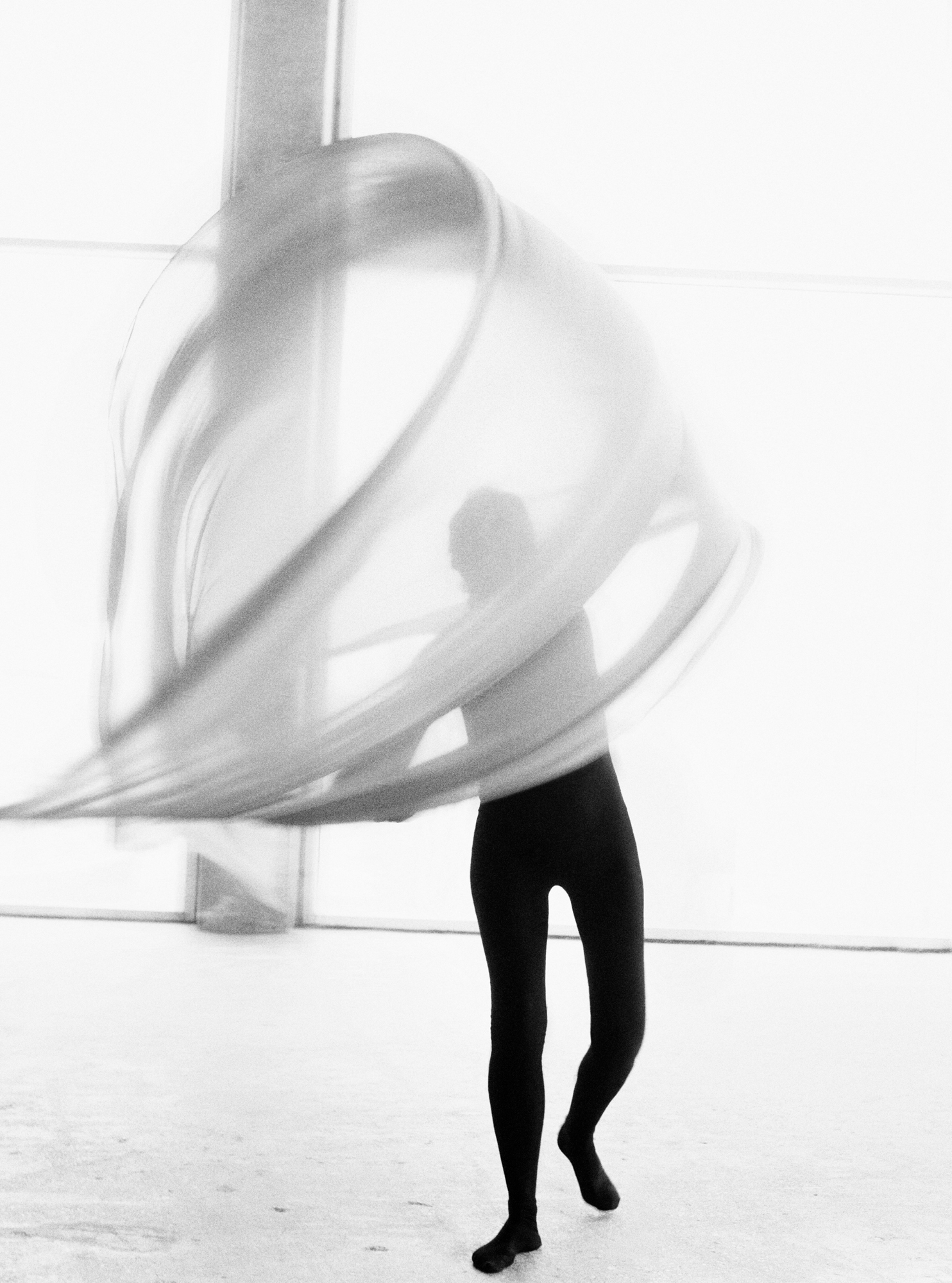A figure is moving a sheer fabric in a circular motion within the frame. The figure is wearing neutral black fitted leggings, and a fitted top. Light is pouring in through the floor to ceiling windows in the background. 