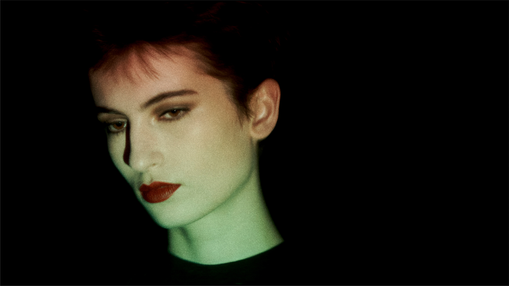 Portrait of a woman with a short haircut against a black background. She has a brown smoked out eyeshadow, and a red lip. The light hitting the model's face starts with green at the neck and jawline, and then fades out at the middle of the face. 