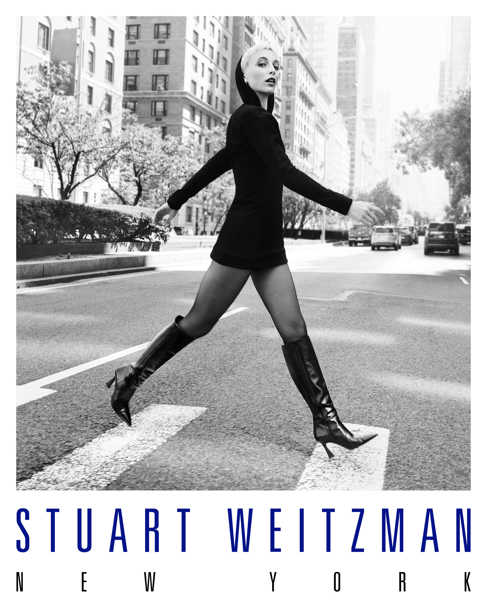 Emma Chamberlain is wearing a hooded long sleeve black look with a mini skirt. She is wearing knee high black leather heeled boots. She is walking confidently across the street on Park Ave in New York City. The Stuart Weitzman New York logo is featured at the bottom of the image. 