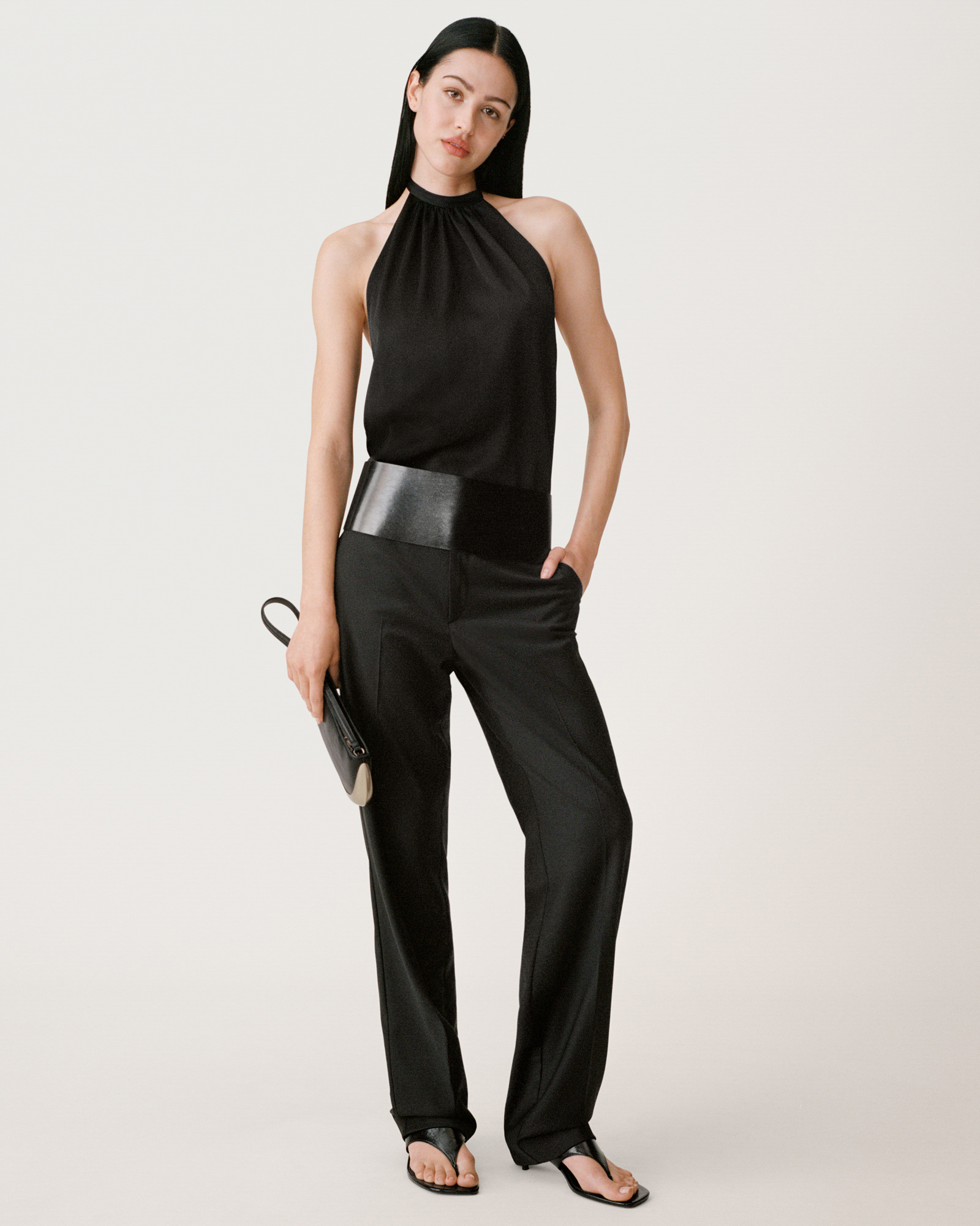 A model is standing in all black against a cream white background. She is wearing a sleeveless top, leather belt, and trousers. She is holding a small clutch bag and has her head leaning down slightly. 