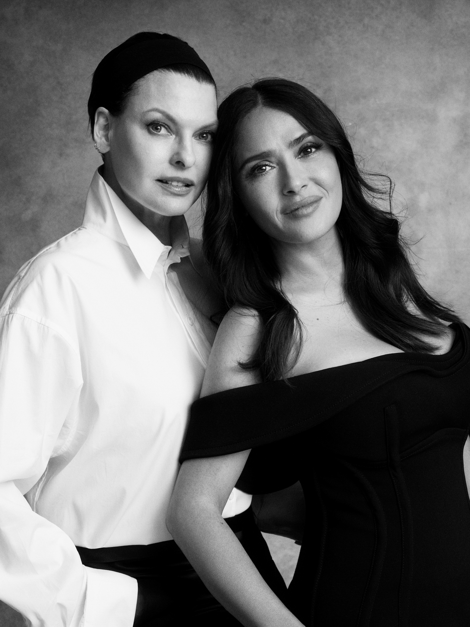 Linda Evangelista and Salma Hayek pose standing next to one another. Salma is leaning her head back towards Linda. Linda is wearing a white button up shirt, and Salma is wearing a shoulder less black dress. 