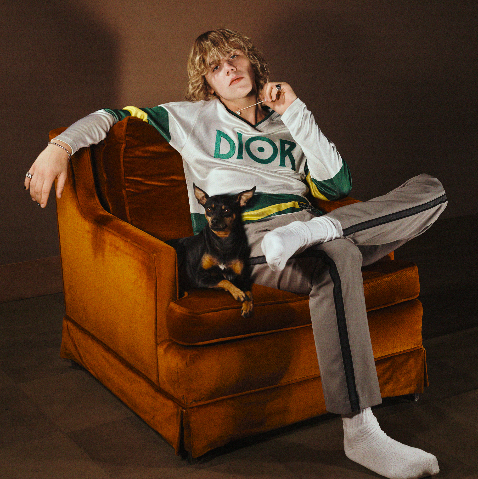 Kid Laroi is sitting in an orange cushioned chair. He has one foot resting on one of his knees. He is wearing a Dior pullover with brown pants, and has a small dog next to him. The background is brown. 