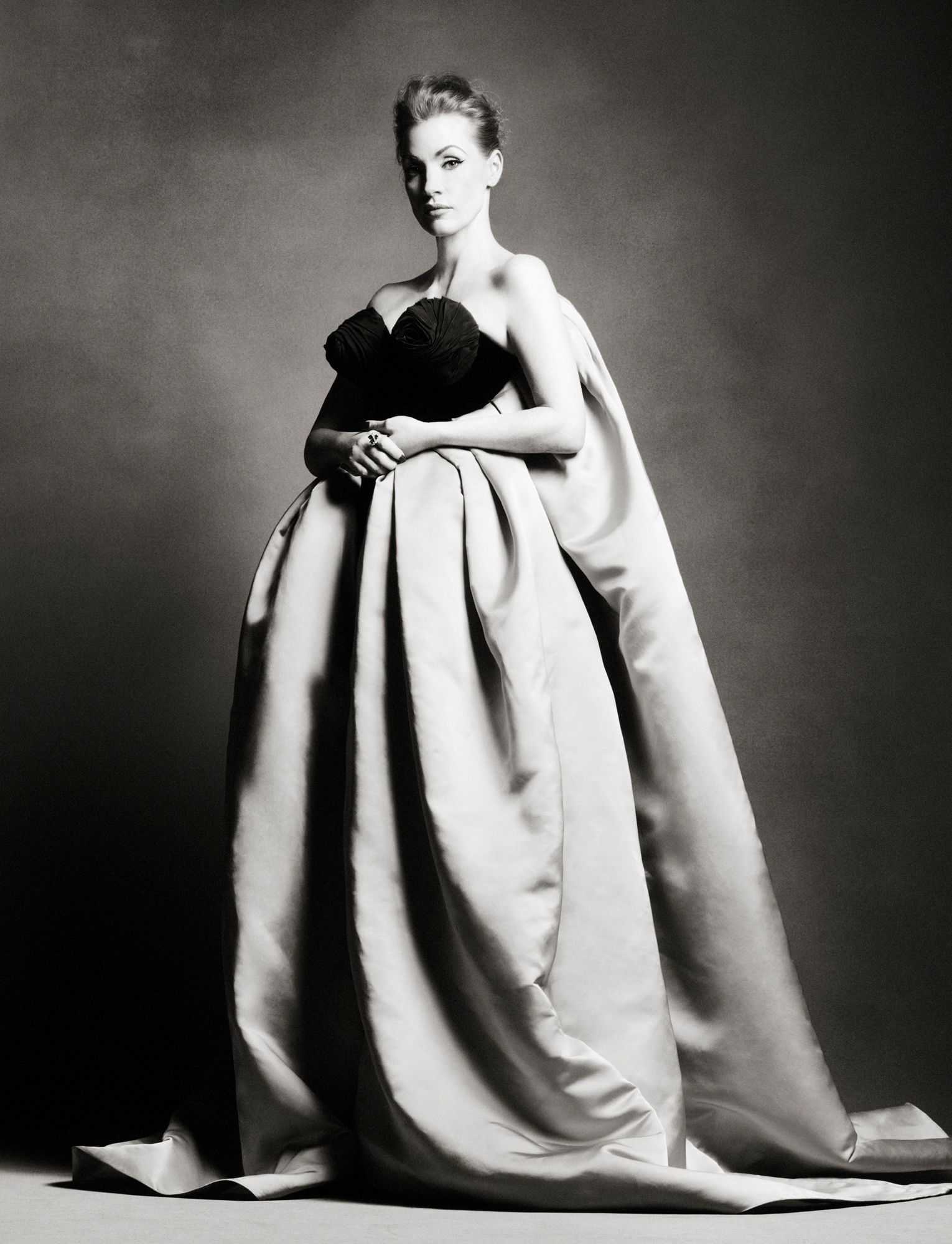 Jessica Chastain is standing in the center of the frame wearing a gown with a black bodice. She is holding her hands together at her waist and is looking directly at the camera. 