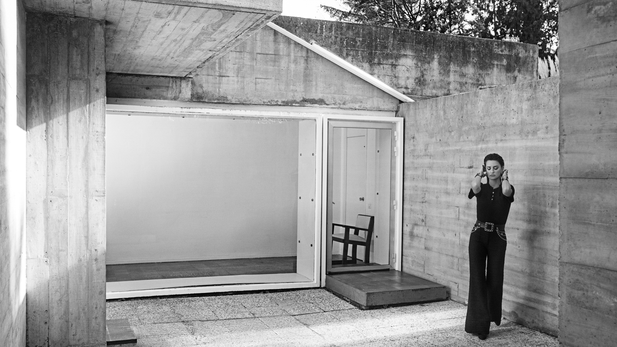 Penelope Cruz is walking forward while putting her hair behind her ears. She is within a concrete courtyard with Brutalist architectural details. 
