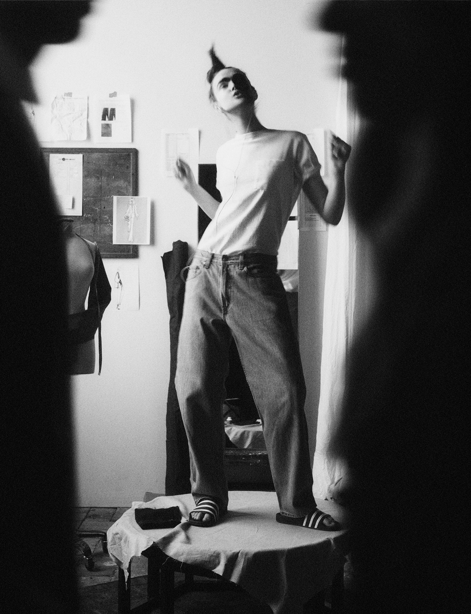 Lulu Tenney is wearing a t-shirt and baggy jeans, she is standing on a platform in the middle of the frame. She has headphones in and appears to be dancing to music. Different fashion drawings and such are visible in the background. 