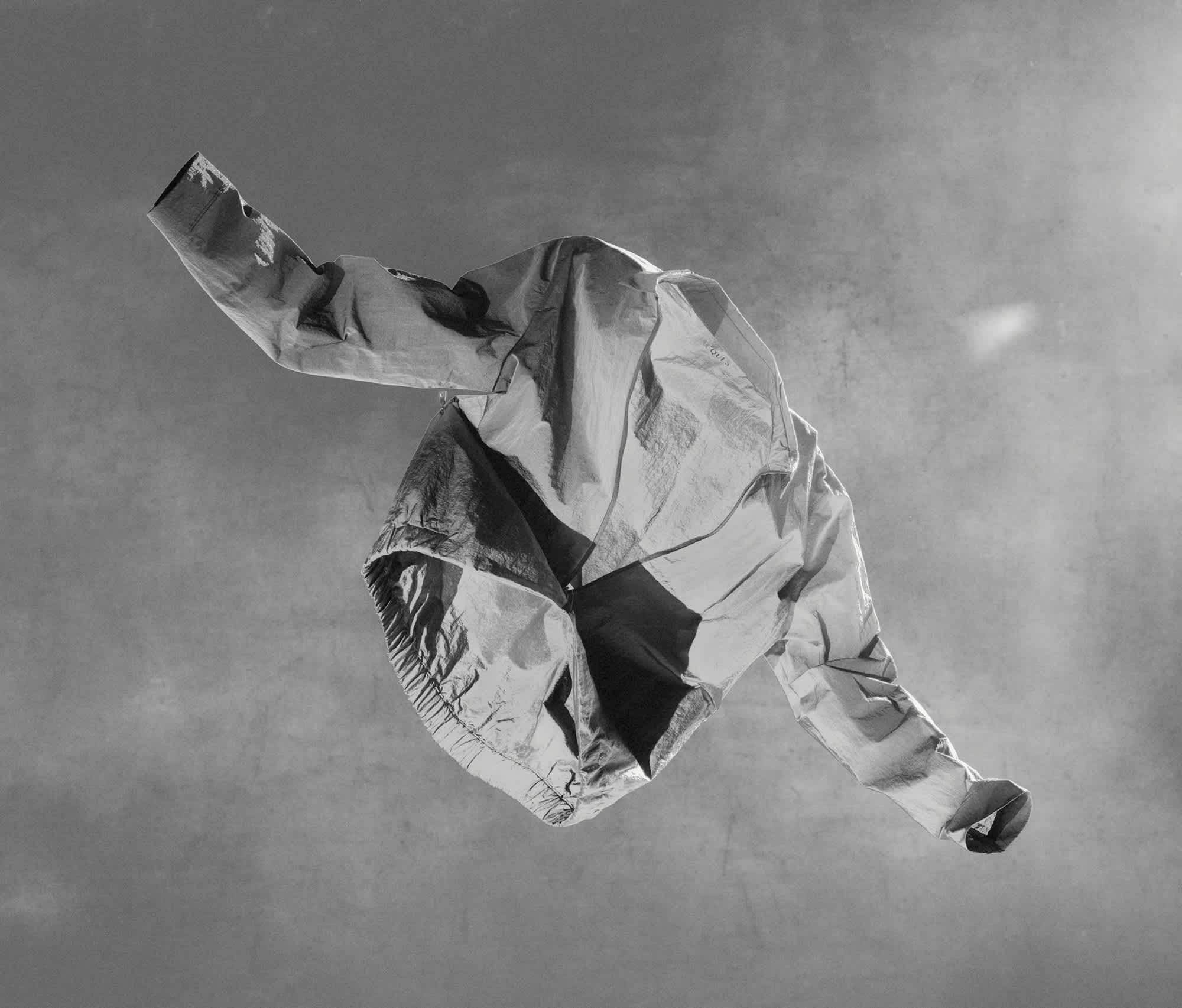 A light jacket has been thrown up into the air, the sleeves extended on the sides. The jacket is mostly unzipped. Light is shining through the jacket, and the background is a textured grey.