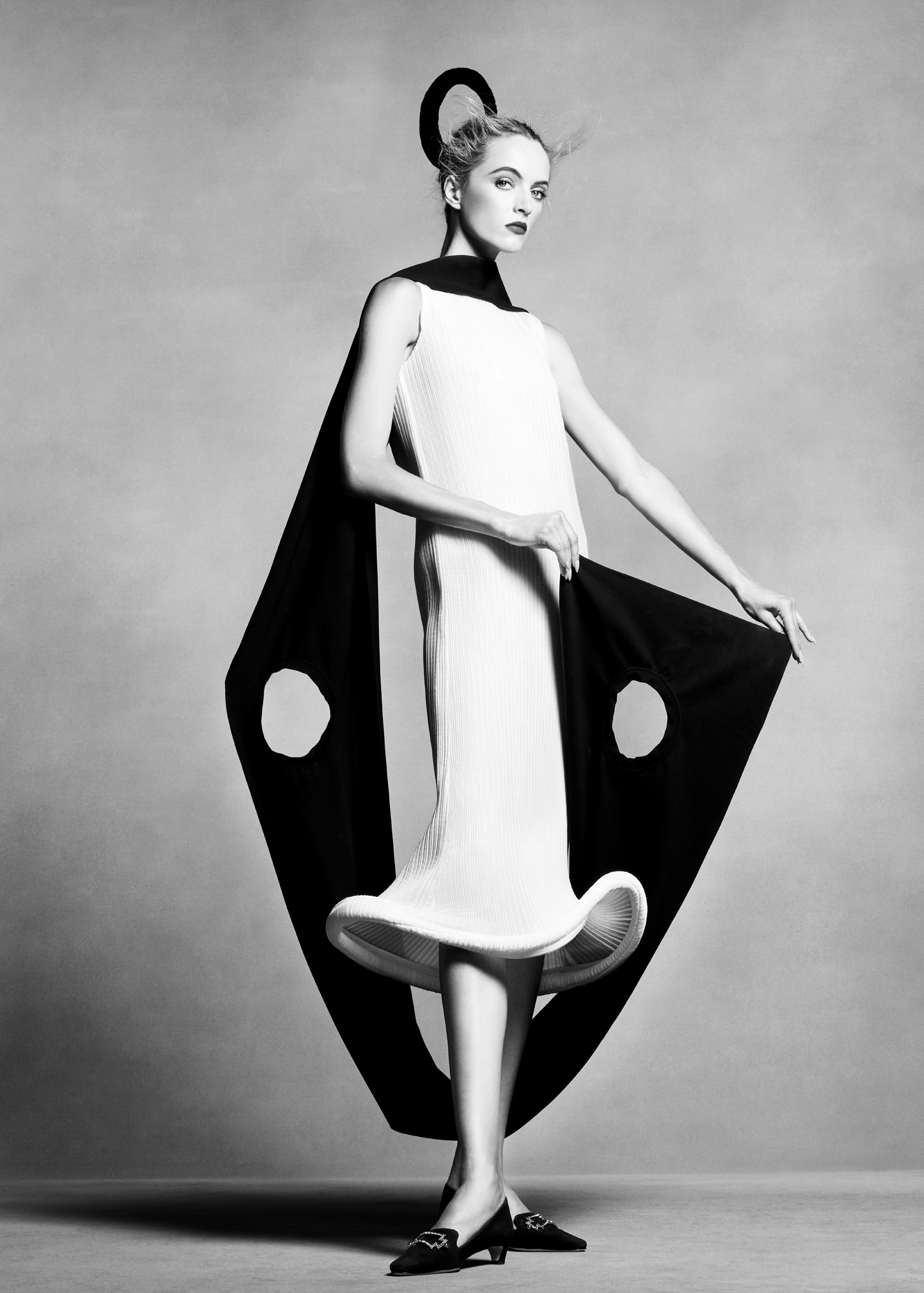 The model is standing in the center of the frame wearing a white dress where the bottom ends in a mushroom like form. She has a cape around her neck that she is holding with both hands forward that has circular cut outs. She has simple black heels on in front of a grey background. 