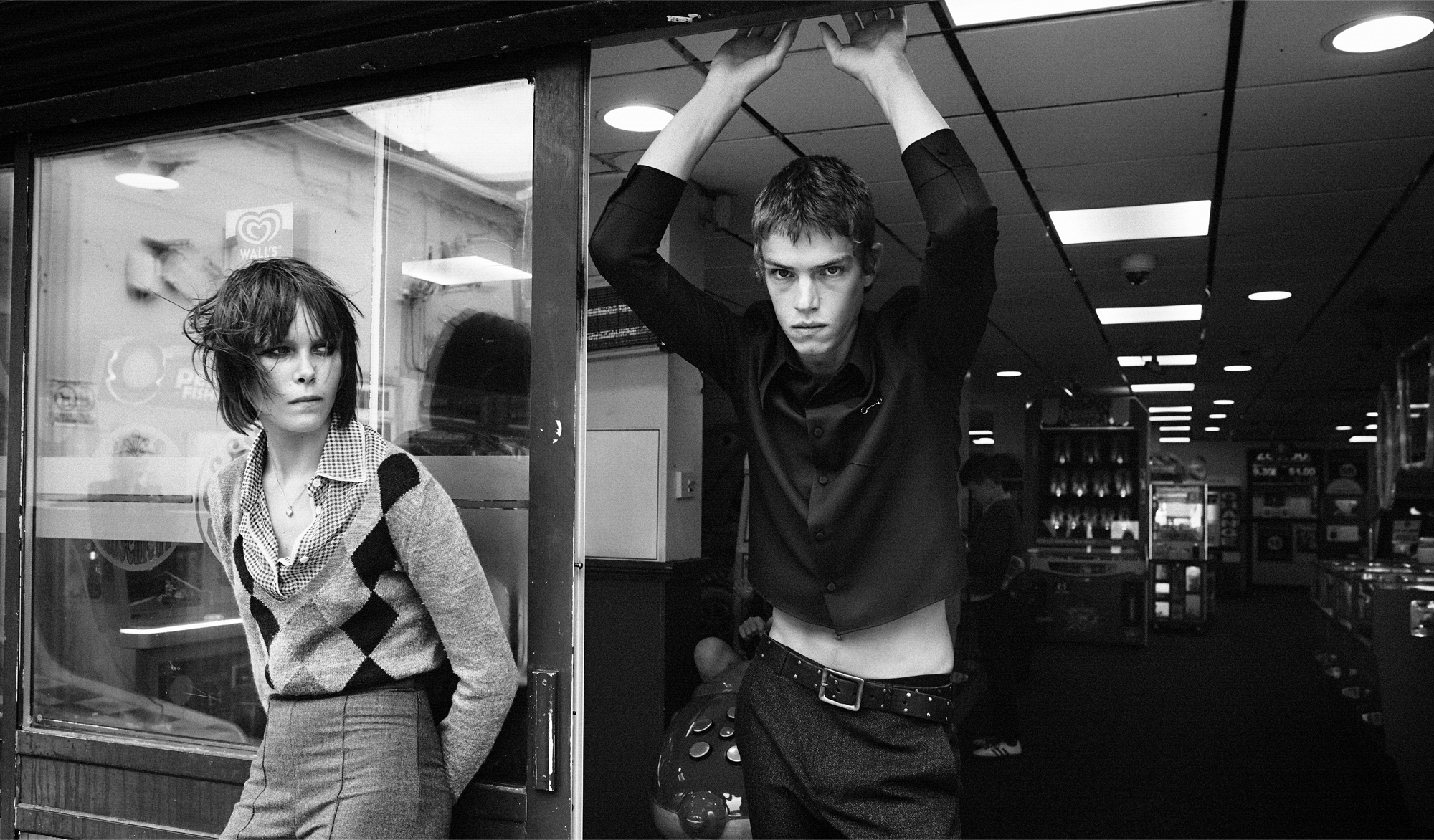 A young man and woman are outside an arcade. The man is resting his hands on the top of the door frame of the entrance. The woman is leaning against a window with her hands behind her. 