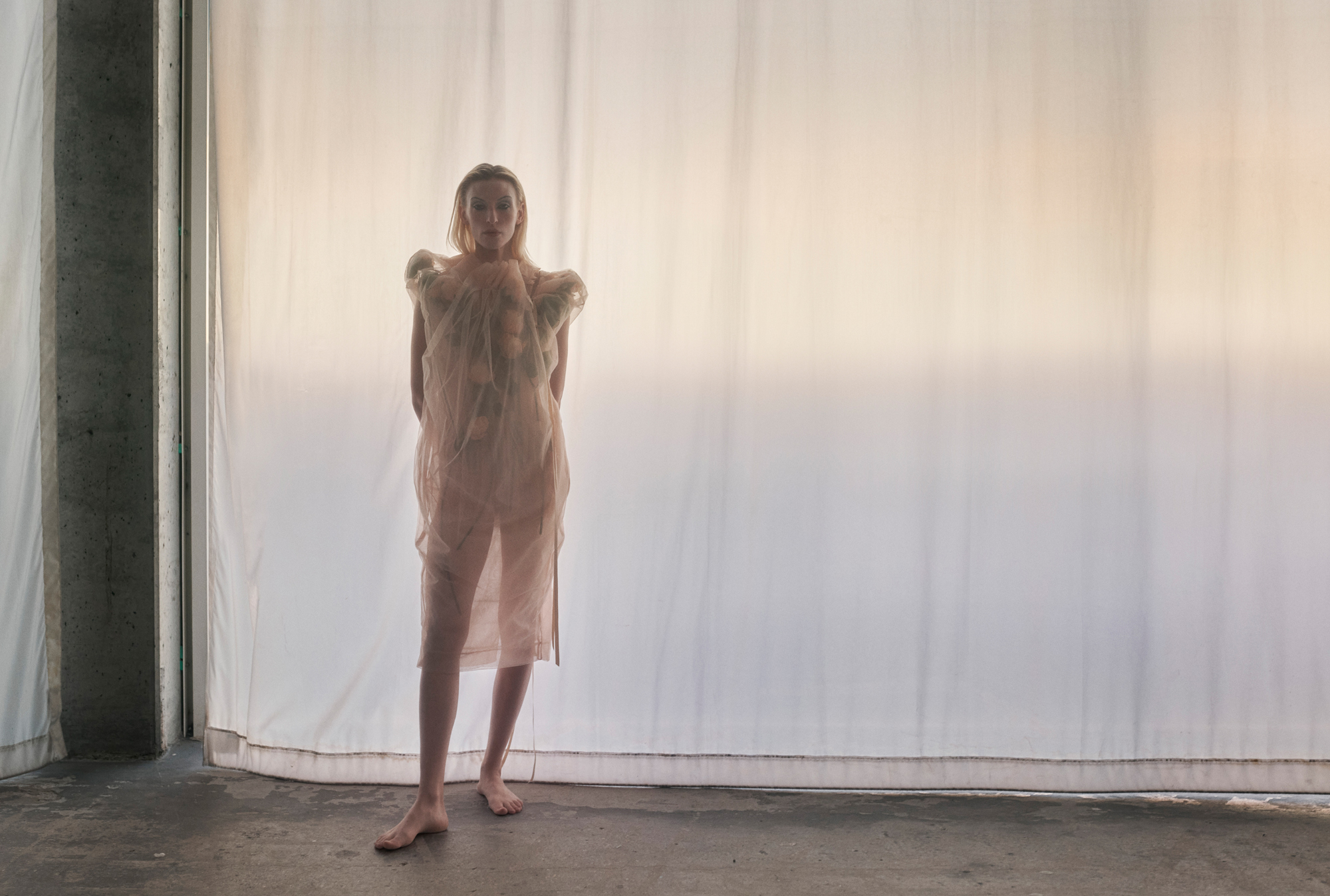 Model is standing towards the left hand side of the image. She is wearing a sheer fabric creme dress. The background is a sheer curtain that is letting through golden hour sunlight. 