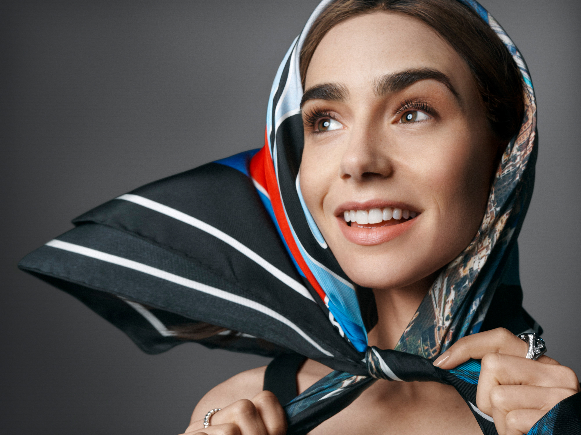 The image is a close up portrait of Lily Collins. She has a multi colored scarf over her head, and is pulling the ends tight with her hands. She is looking off to the left past the camera. 