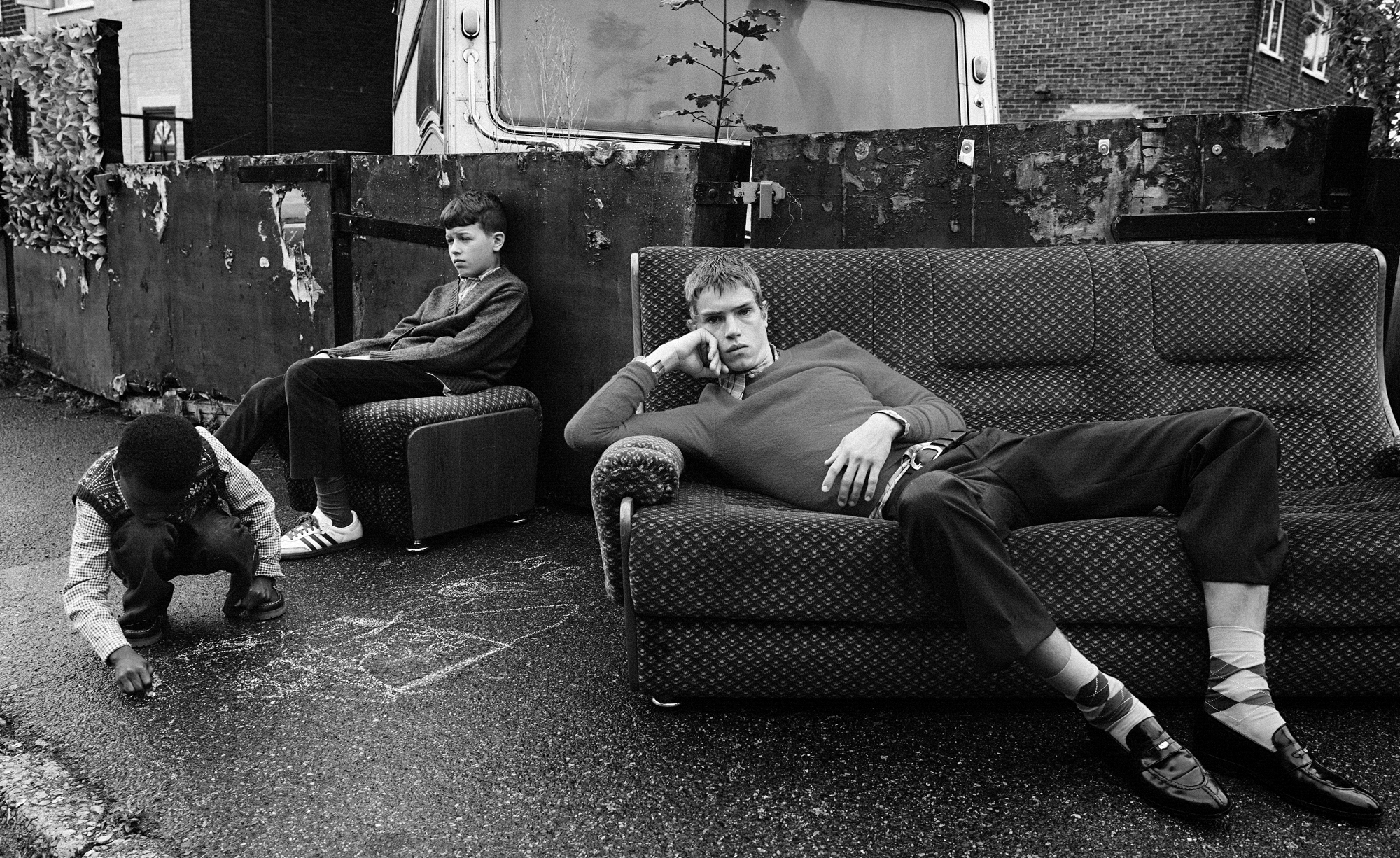 A young man is reclining on a sofa that is outside on the sidewalk. A young boy is seated on a piece of furniture while another boy is playing with chalk on the sidewalk. 