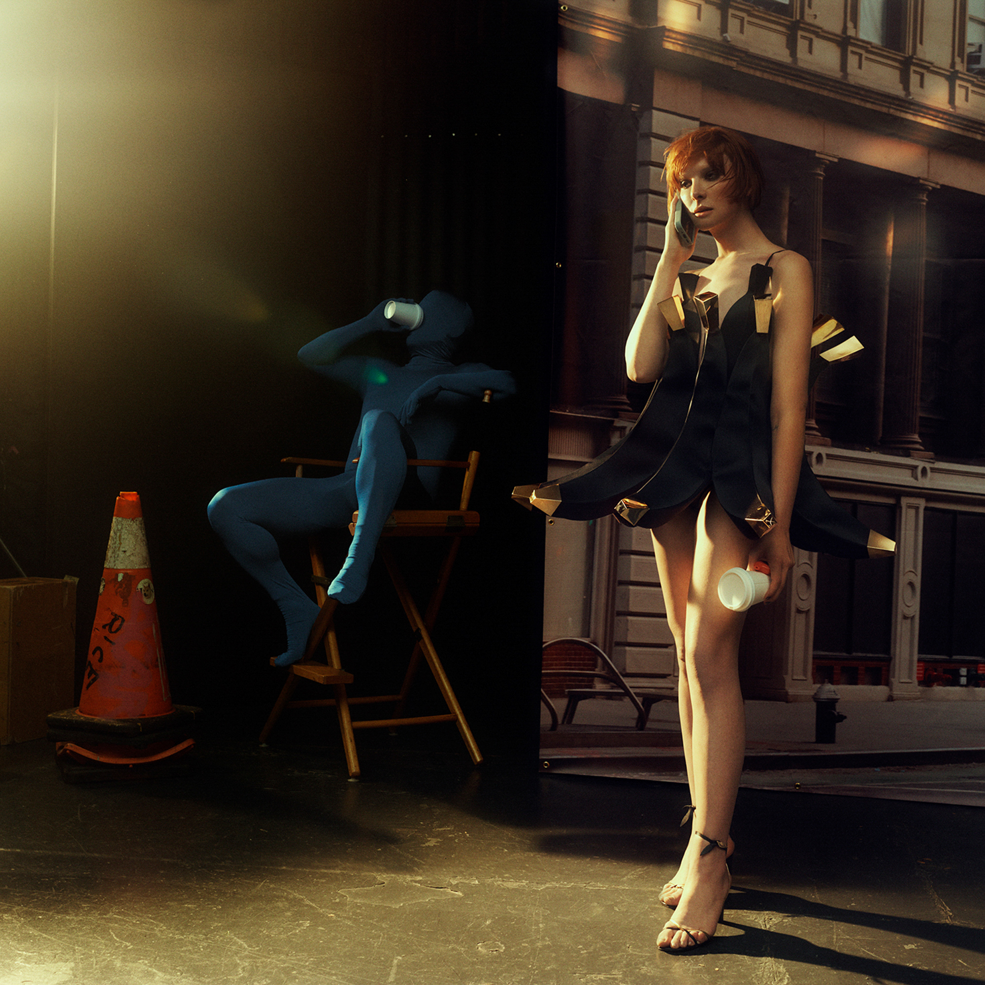 Hari Nef is wearing a banana dress and heels, she is holding a phone up to her ear and holding a to go coffee cup. There is a male figure in a head to toe form fitting blue suit drinking from a to go cup of coffee. He is seated in a directors chair. There is a caution cone in the left hand corner of the image, light coming in from the top left corner, and a New York City building backdrop that cuts off right at the middle of the image. 