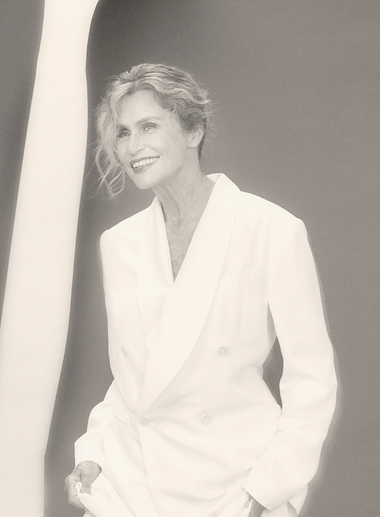 Lauren Hutton is smiling looking off to the right of the camera. She is wearing an all white suit standing in front of a grey background with a white strip that is on the left hand side of the frame. 