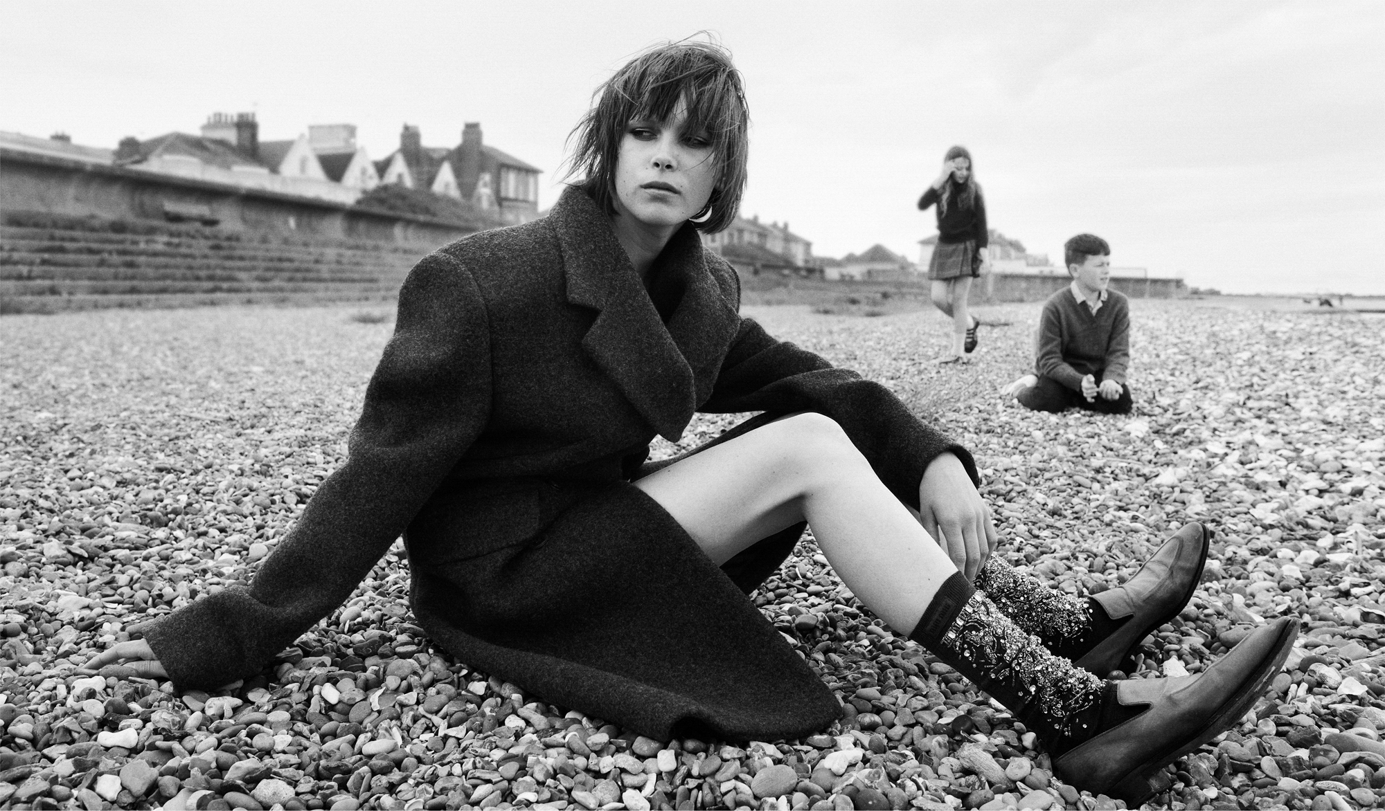 A woman wearing a knee length coat is seated on a rocky beach looking off into the distance towards the left hand side of the frame. Two children are visible on the beach behind her. 