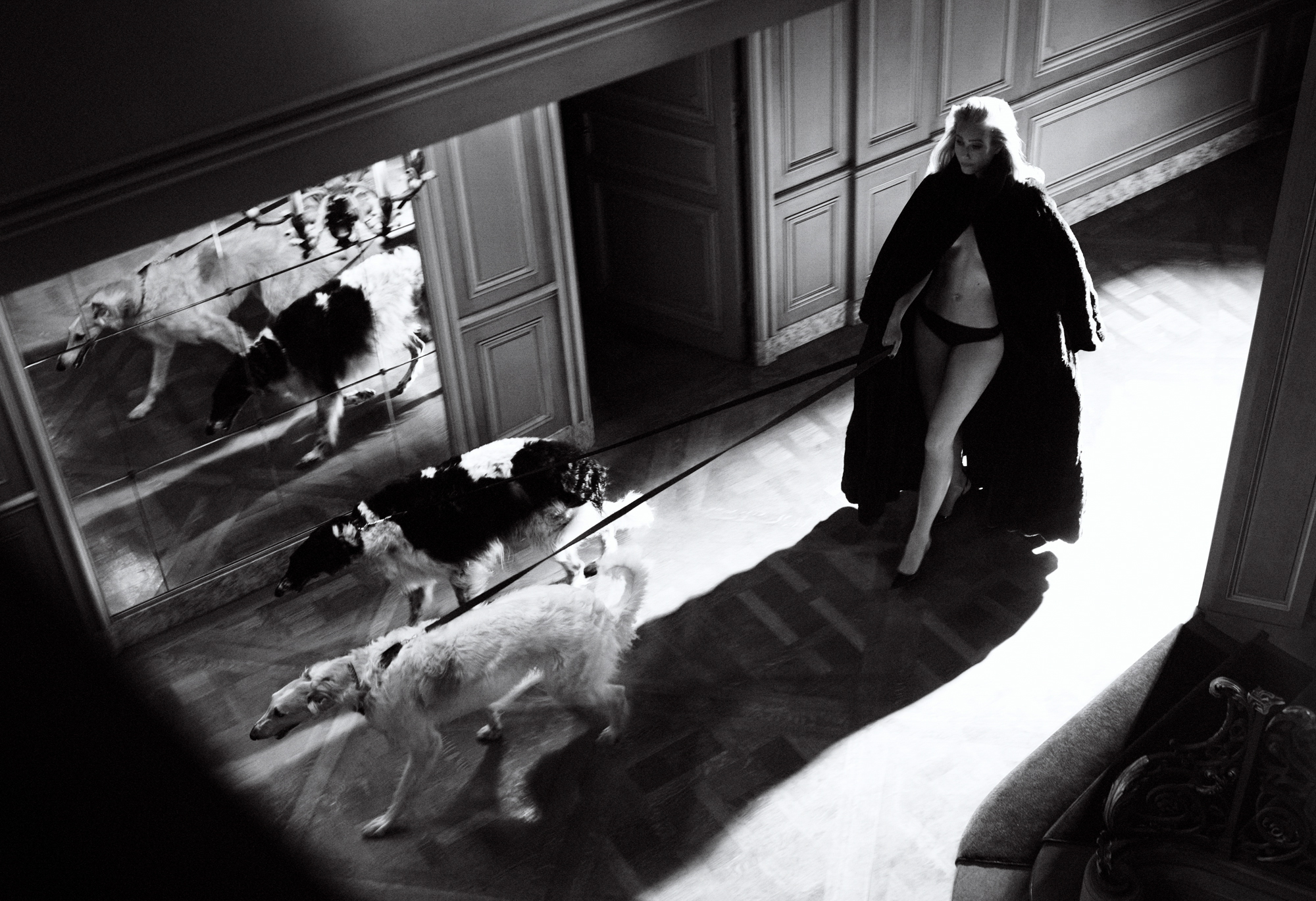 Gwyneth Paltrow is wearing a large coat with only underwear on underneath. There are two dogs on leash walking ahead of her in a residential hallway. 