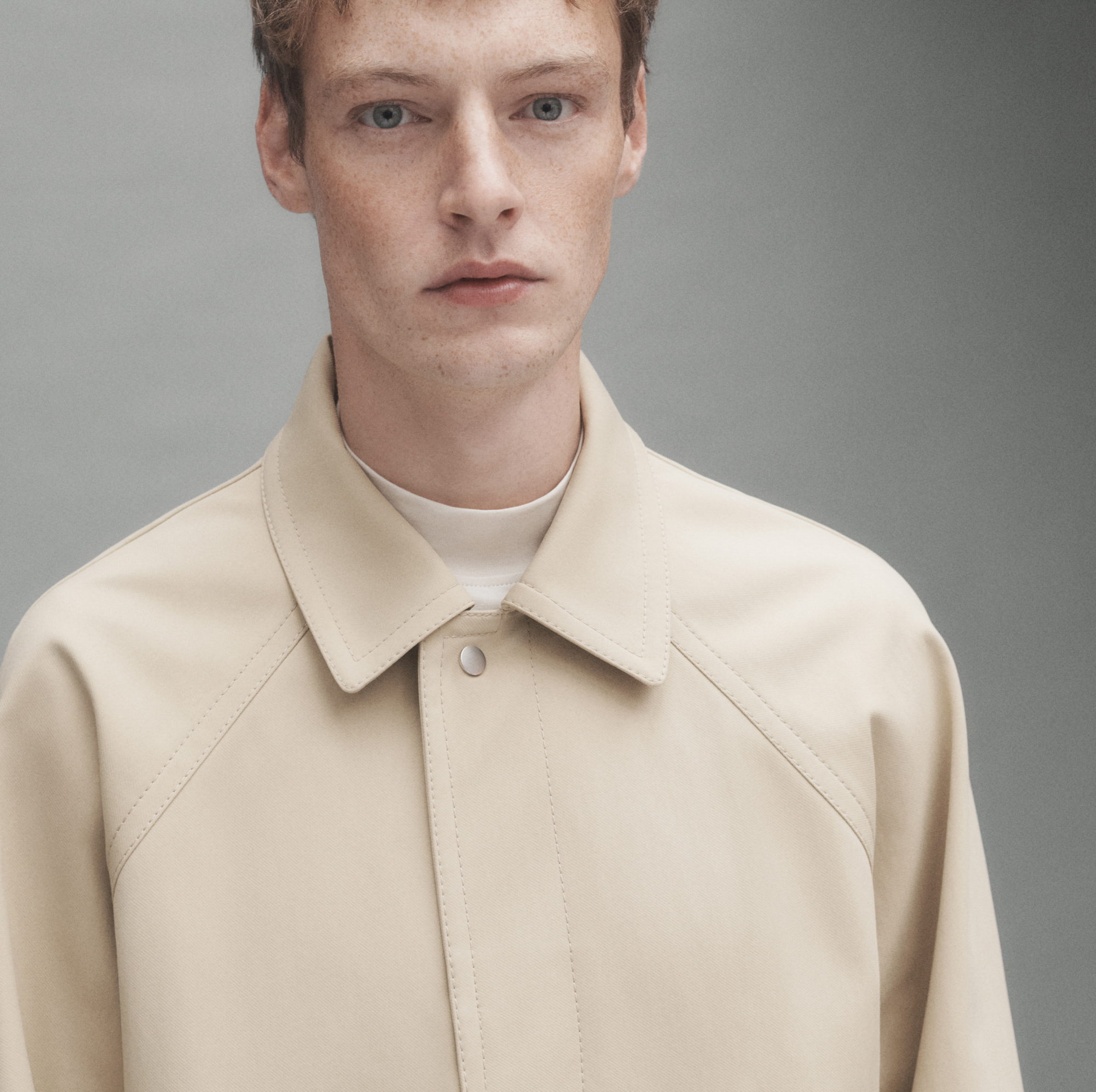 A man with freckles, reddish blonde hair, and blue eyes is off center in the frame. He is visible from the mid torso up, and is wearing a beige trench coat with a collar and silver button on the top. A white mock neck shirt is visible underneath. The background is a grey that is darker from the right and gets lighter as it moves to the left. 