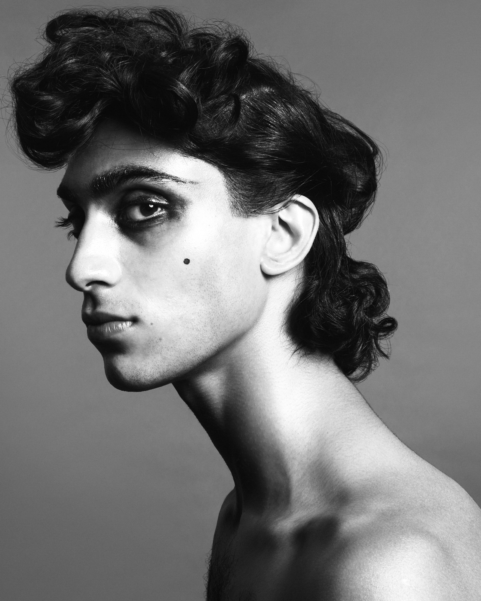 A male model is in the frame and seen from the side. They are visible from the shoulders up, have smoky shadow applied to their eyes, false mole, and sculpted curls that are grouped on the top of the head and on the neck. 