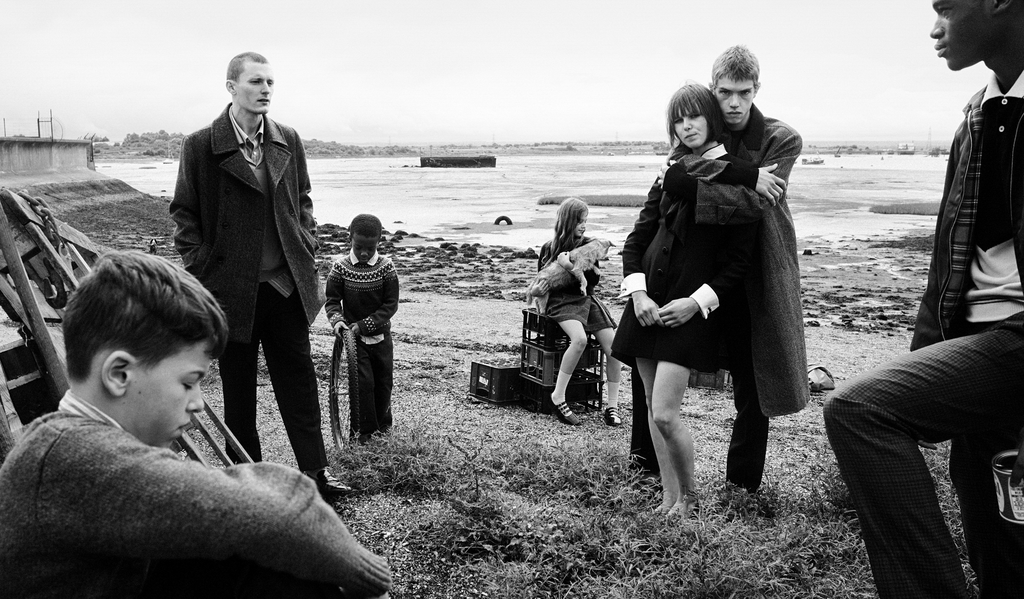 A crowd of people of various ages and ethnicities are gathered on a rocky beach with the sea in the distance. 