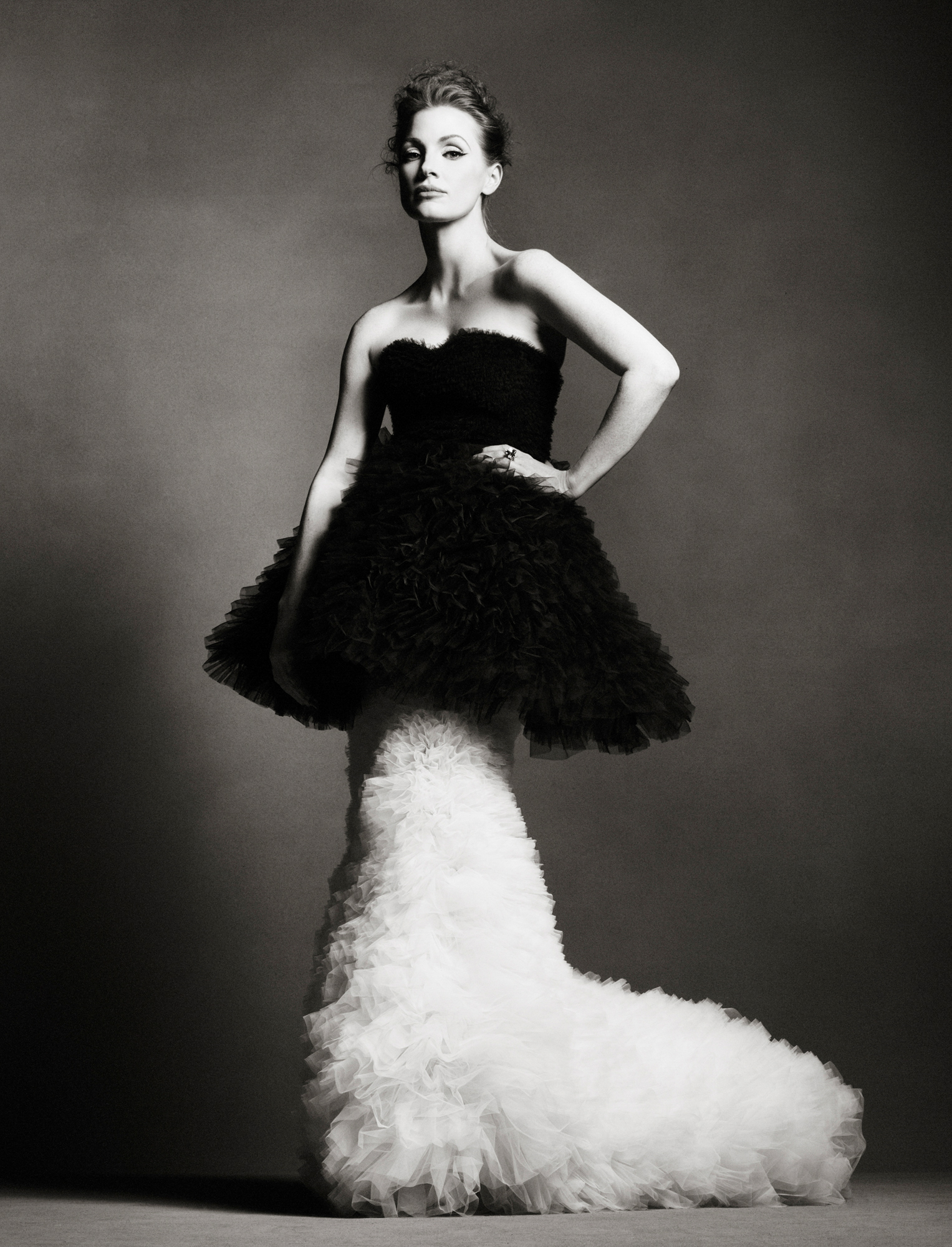 Jessica Chastain is standing in the center of the frame with her hand on her hip. She is wearing a frilly tulle gown, where the top half is black, and the bottom half is white. Background is a textured grey. 