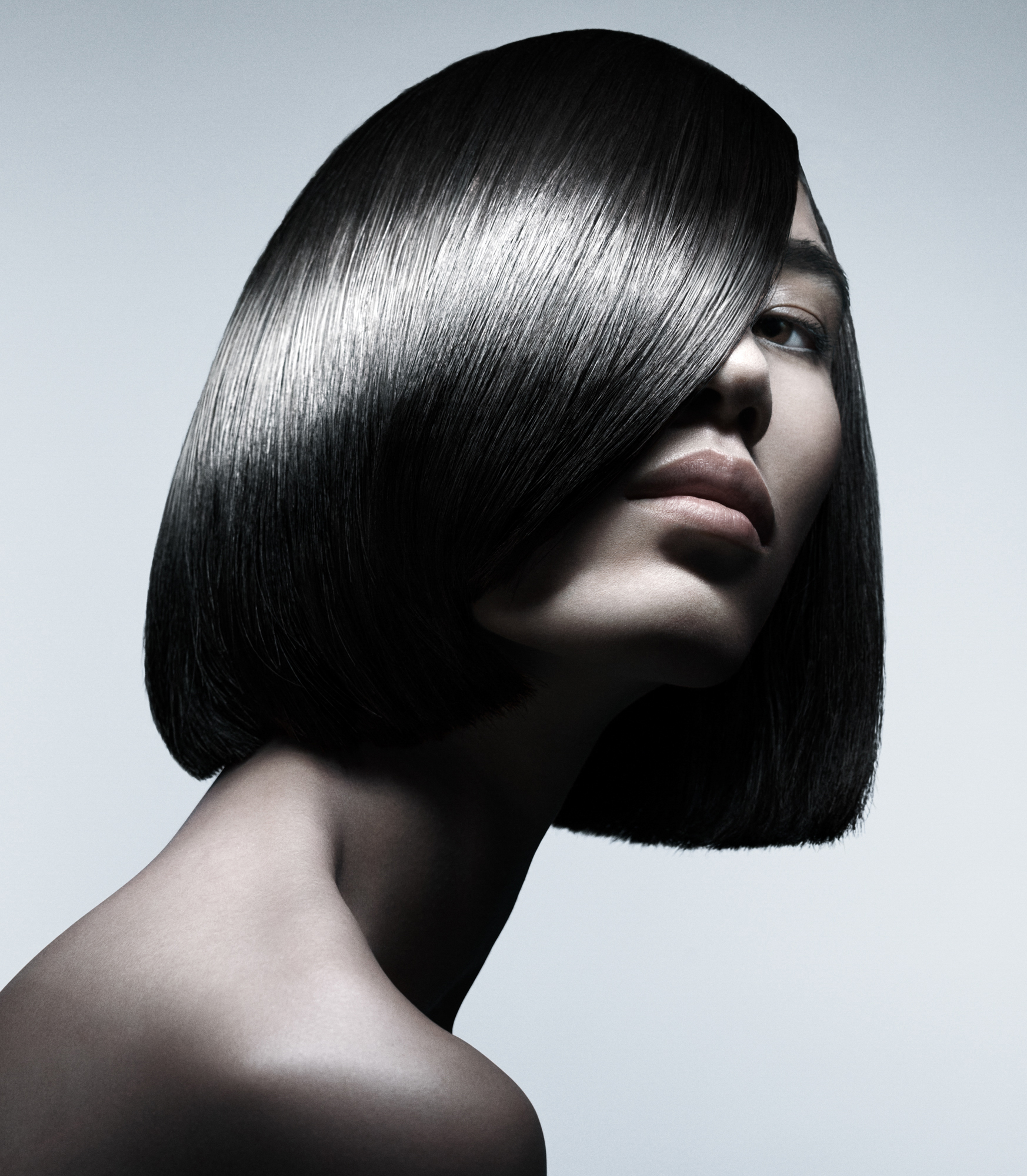 A woman with sleek black hair has her face partially covered by her hair. She is peering out behind her hair and looking directly at the camera. She is seen from the shoulders up against a blue / grey background. A beam of light is illuminating the shine of the hair towards the center. 
