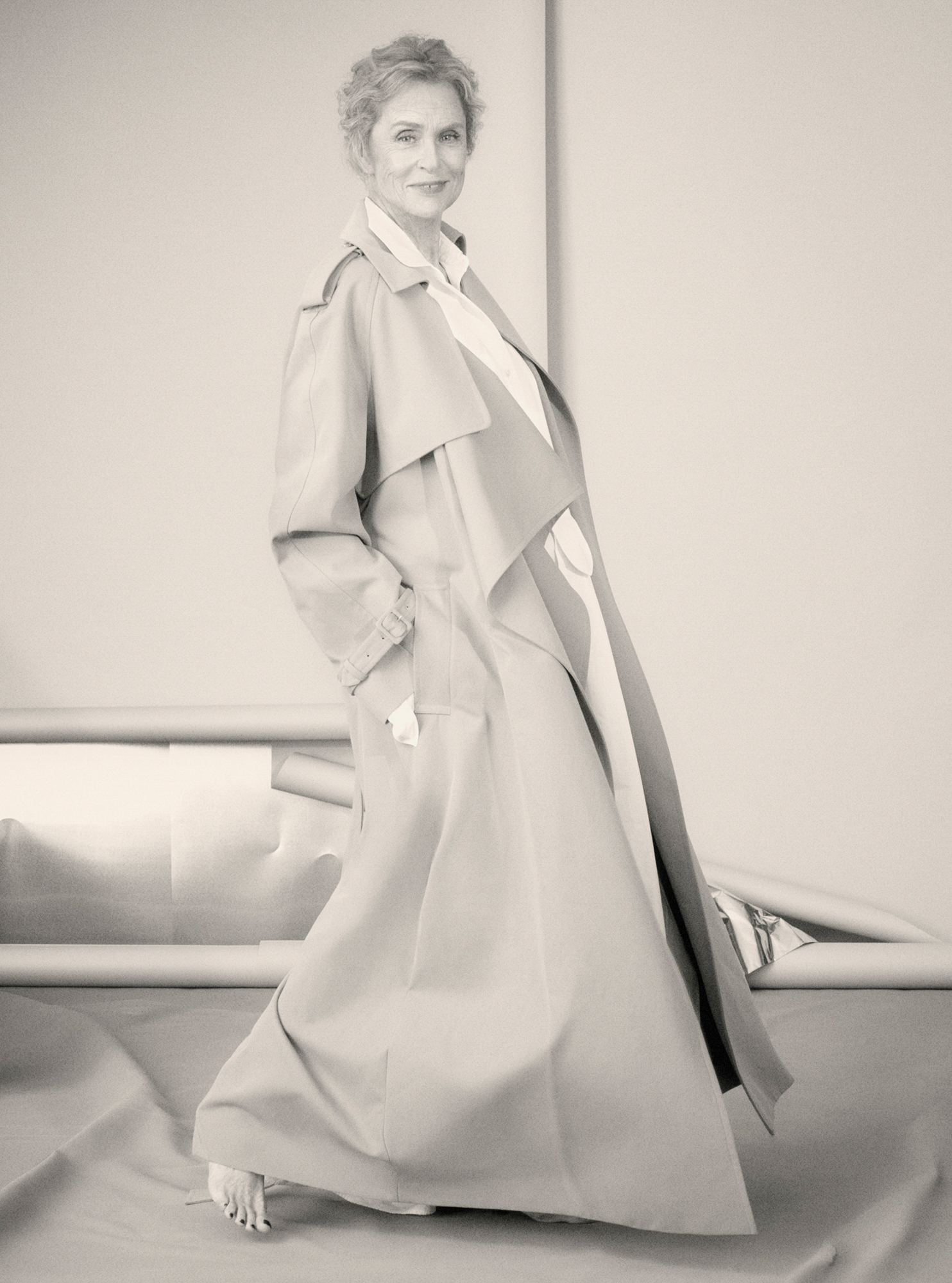 Lauren Hutton is seen with short hair with a floor length trench coat and is barefoot. In the background there are various rolls of set background paper. 
