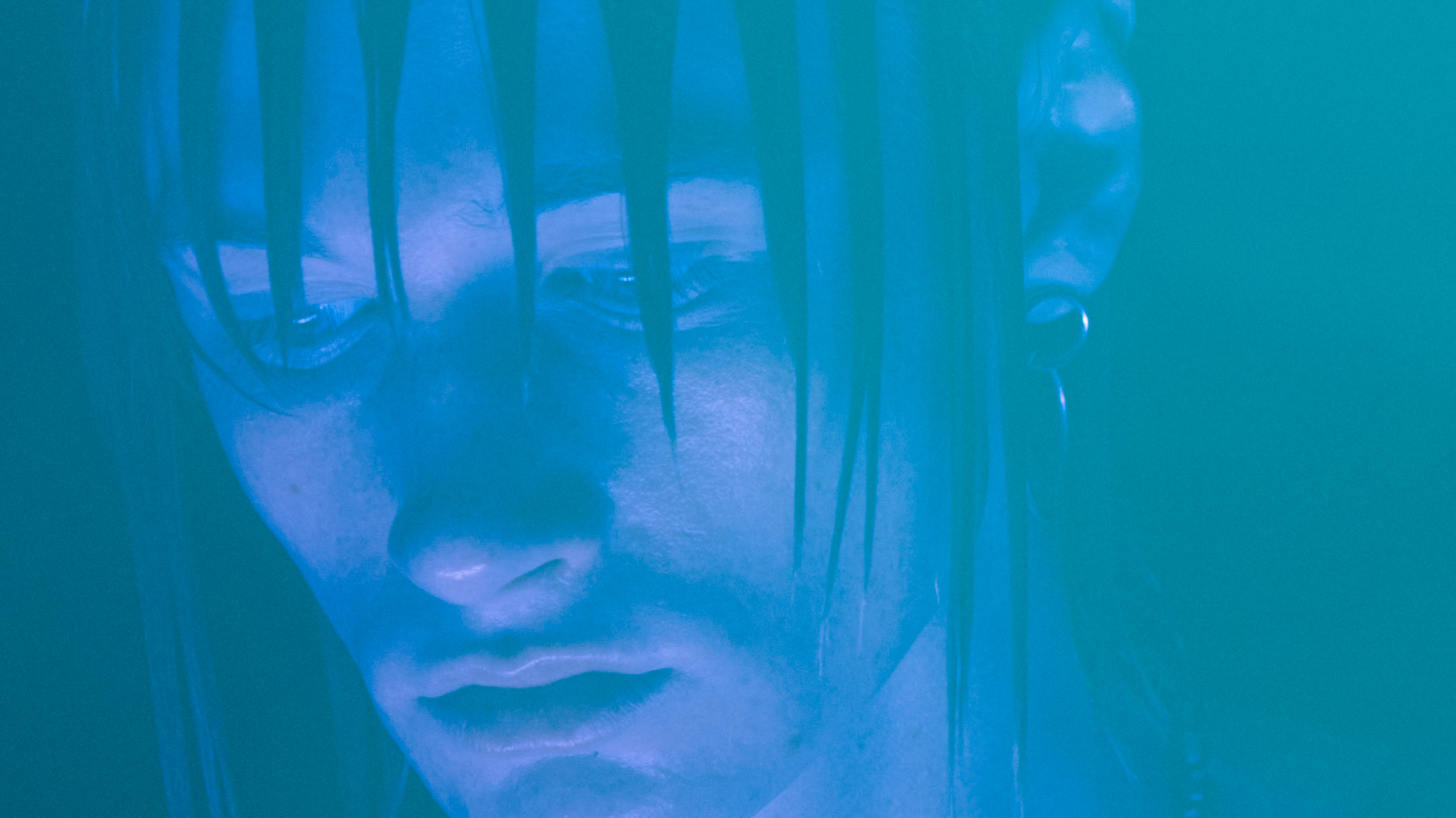 A male model is looking down at a purple toned light. He has an earring and dramatic spiked hair that is coming down over parts of the face. 