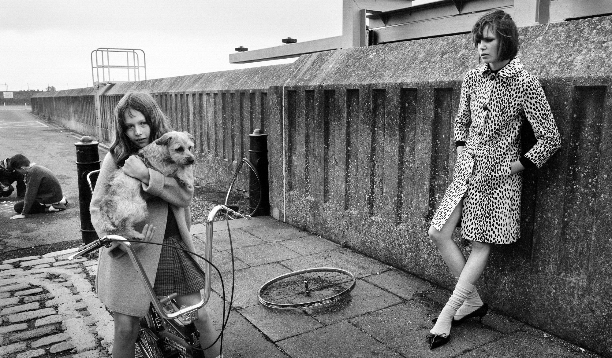 A woman wearing an animal print coat is leaning against a stone wall while a young girl holds a dog while on a bike. Other children can be seen in the distance playing on the ground. 
