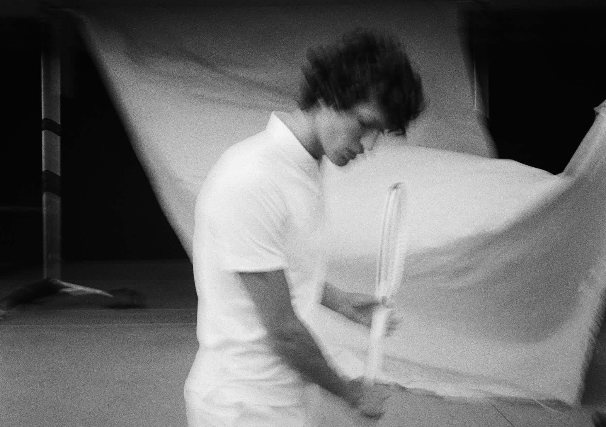 In the frame there is a man with dark curly hair that has his head tilted down, and has a tennis racket in both hands. He is wearing a white shirt, and in the background there is a fabric that the blowing towards the camera. The entire image is slightly blurred.