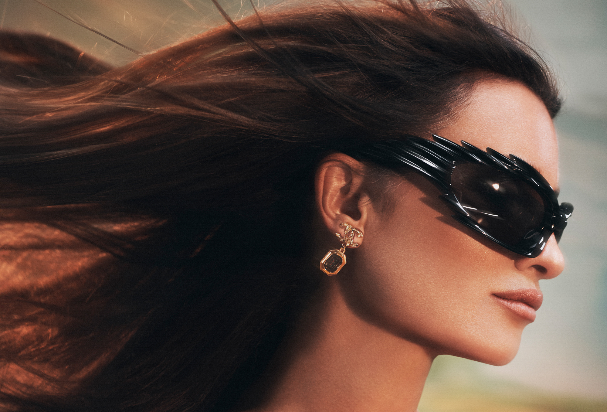 Close up shot of Penelope Cruz. She is wearing large black sunglasses with small spike edges on the frames of the glasses. She has Chanel earrings on. Her hair is blowing behind her. 