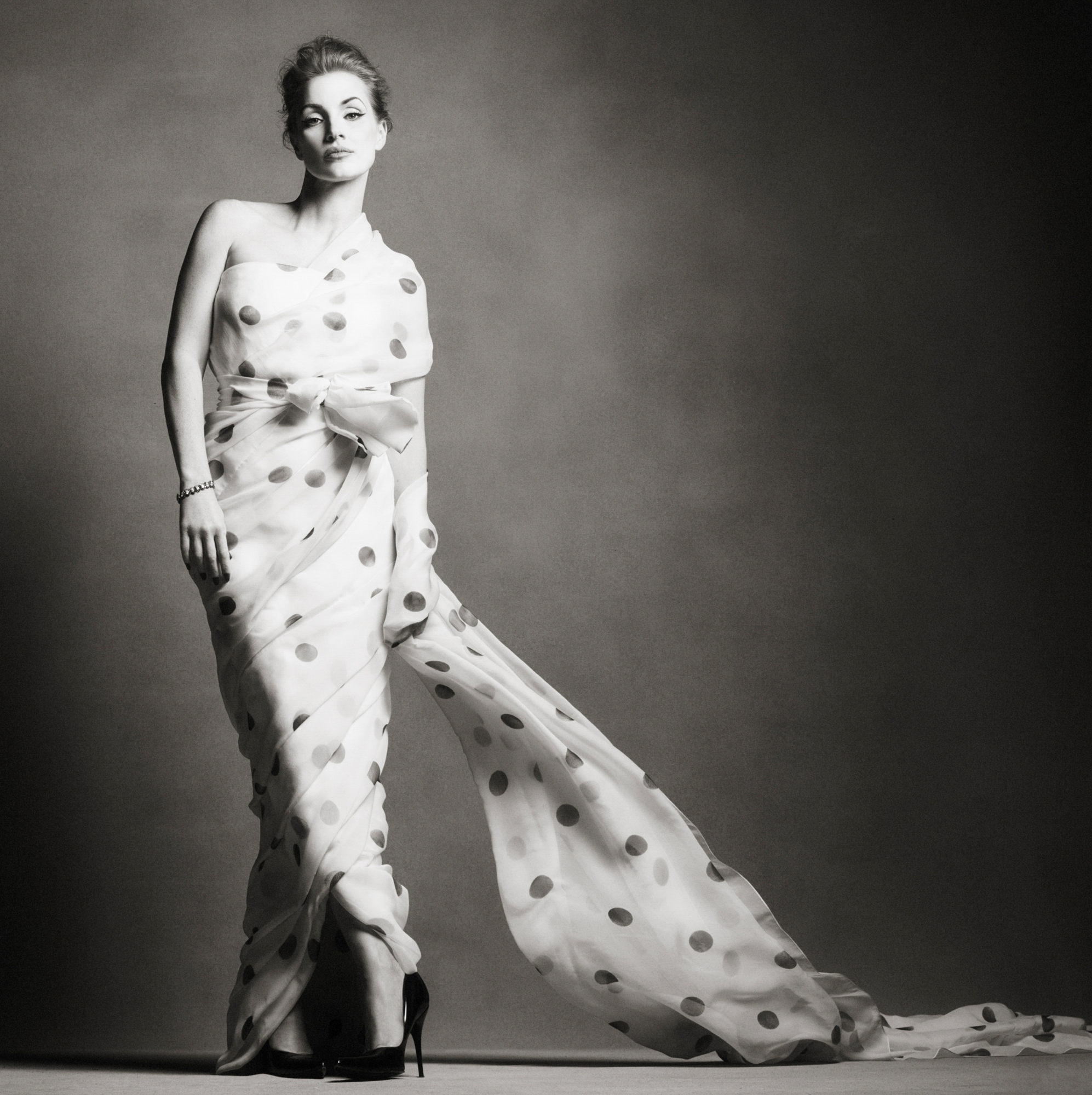 Jessica Chastain is standing towards the left of the frame. She is wearing a polka dot dress with a train that is hanging off the dress towards the right hand side of the frame. She is standing in front of a textured grey background. 