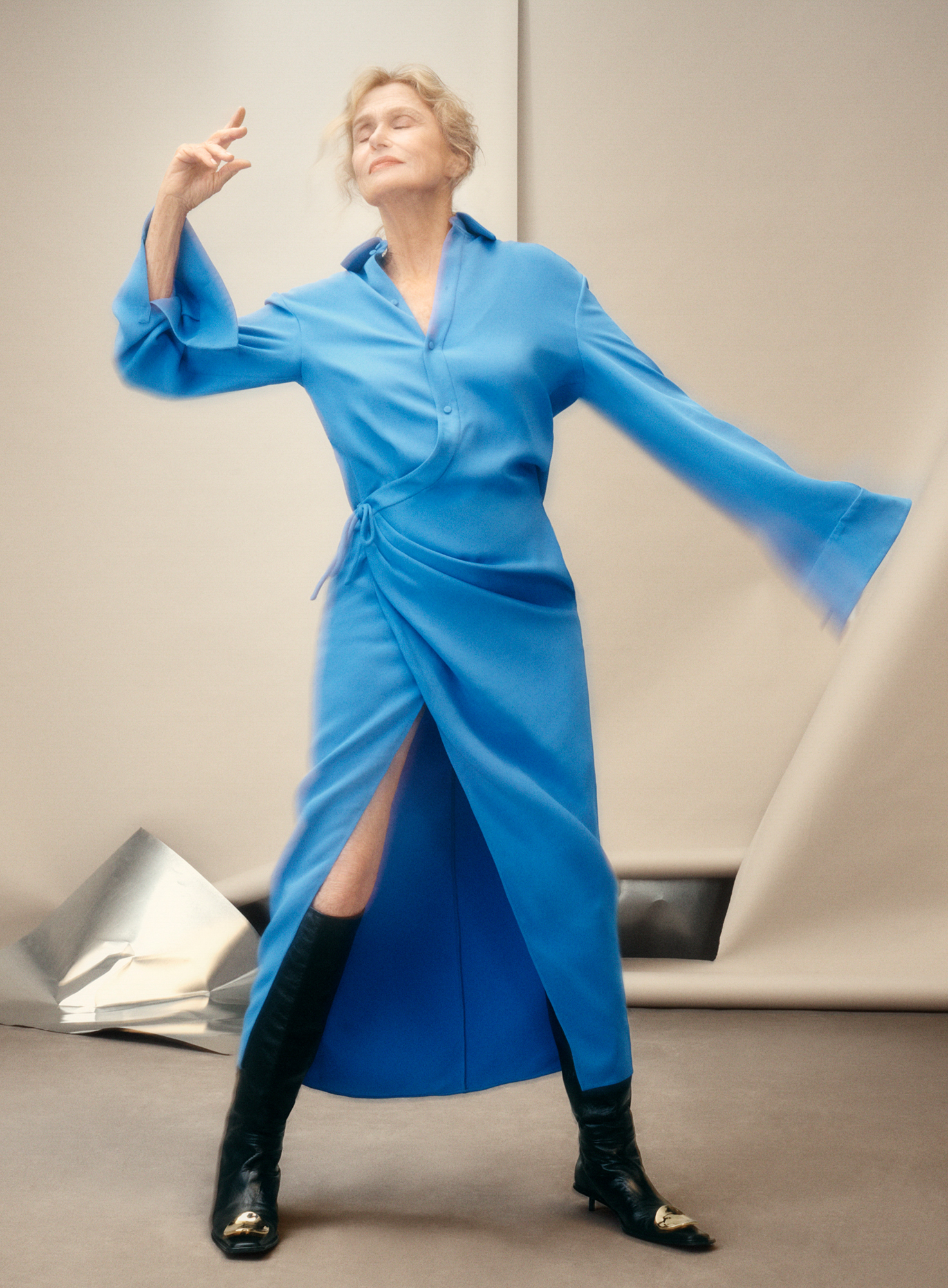 Lauren Hutton is standing a blue dress with knee high black boots. She has one hand pointed up to the ceiling, and has her eyes closed and head tilted slightly back. There is motion blur on sections of her figure. 