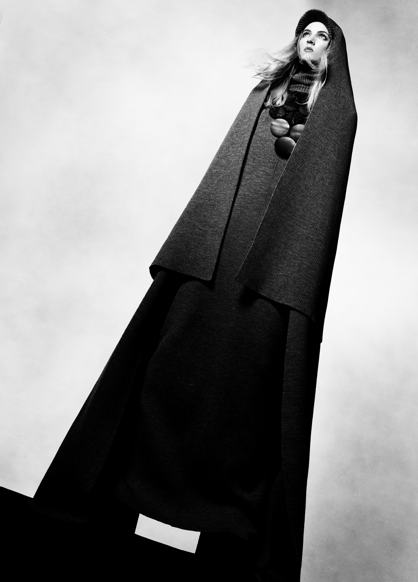 The model is standing at a diagonal, wearing a full length dress and a cape that covers the head over it. There is a large circular necklace on her chest. The shot is taken from below, and the perspective on the image makes the model appear gigantic. 