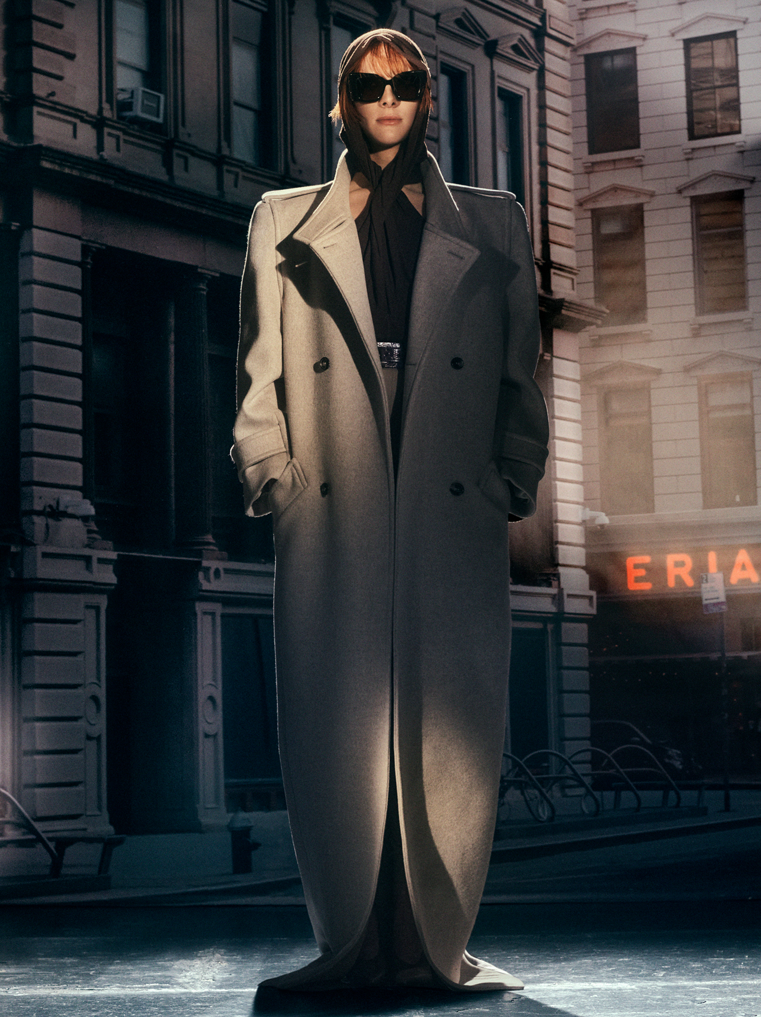 Hari Nef is standing in the center of the frame, she is wearing a full length trench coat that touches the floor. She has a hood on and is wearing large square black sunglasses. A warm spotlight is illuminating sections of Hari's figure. In the background there is a backdrop of New York City buildings. 