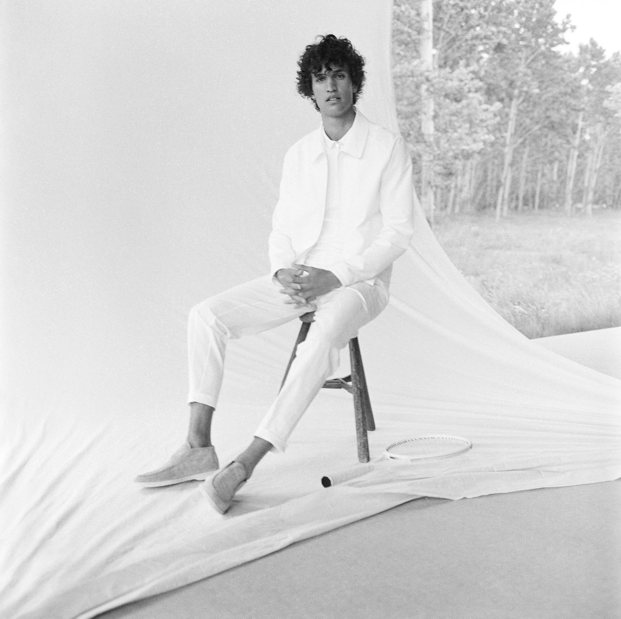 A man wearing all white is seated on a wooden stool. He is wearing a jacket, shirt underneath, and pants. There is a tennis racket on the ground next to the wooden stool. There is a white fabric that is being pulled forward as a background, to the right of the frame the woods behind the scene are visible. There is another grey surface on the floor of the scene. 