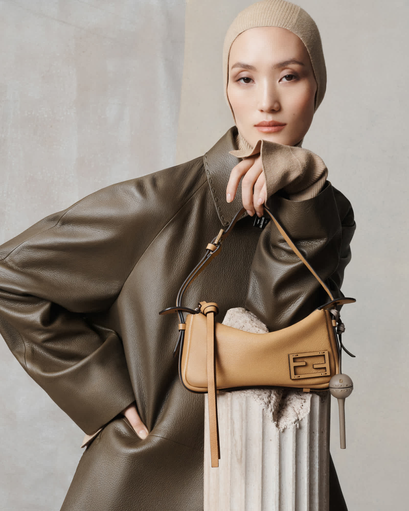 A woman is wearing a brown leather coat and a light brown fabric head covering. She is resting her elbow on the top of a Greek column, and on the column a light brown Fendi bag is resting on the top. The backdrop is two different grays, one with more texture, and one with a more even surface.