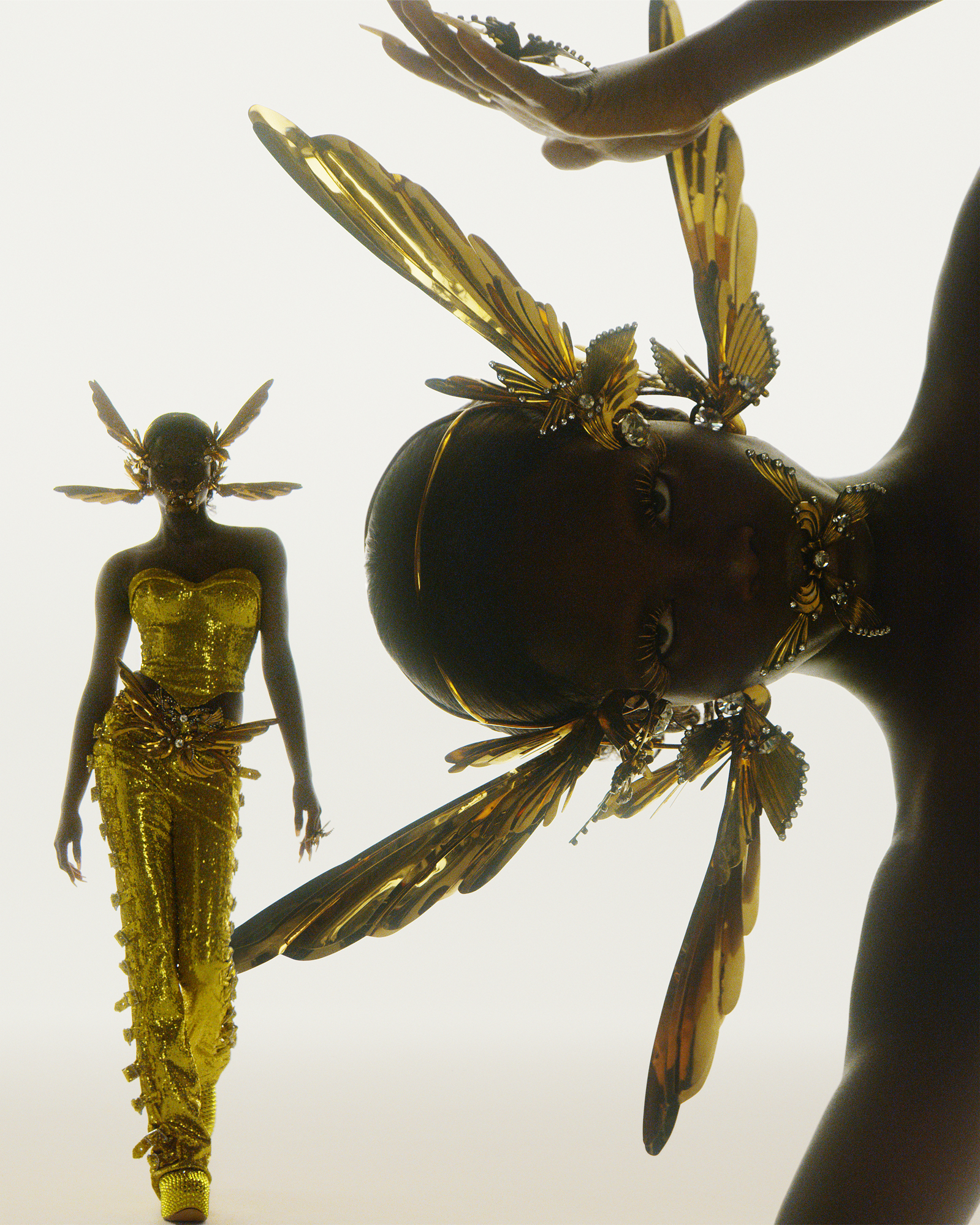 There are two figures within the frame. One is seen on the right side from the shoulders up, and is wearing a headpiece with insect looking wings on it. The figure on the left is seen from head to toe, and is wearing a yellow dress with the headpiece with insect looking wings. 