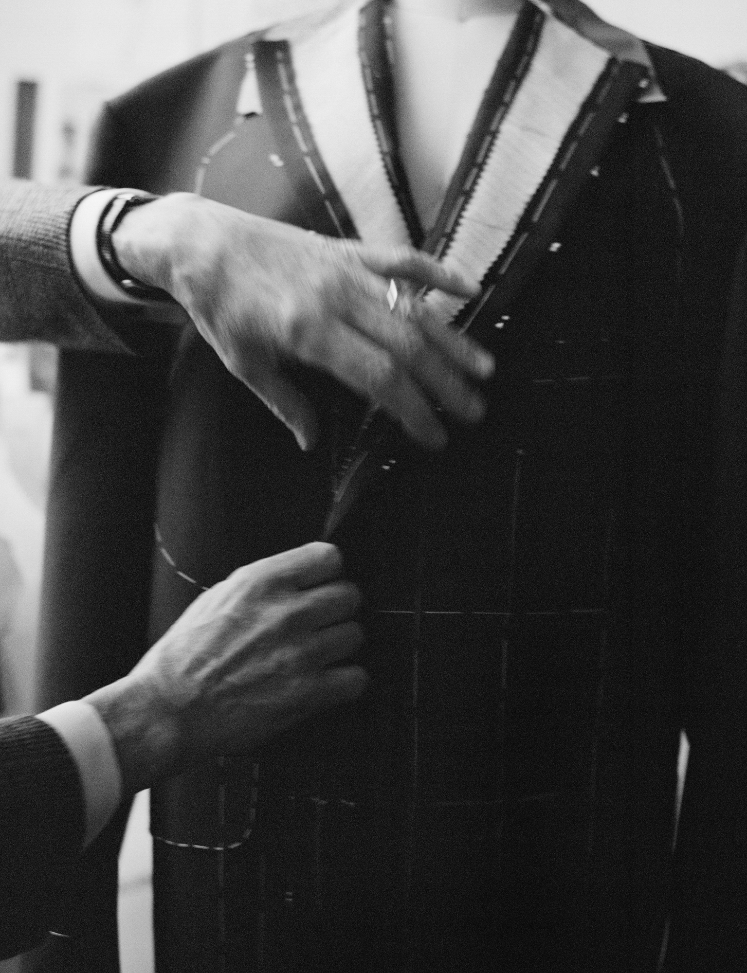 Hands of an older man come into the frame from the left hand side to adjust the middle of a jacket. The jacket is unfinished, and is on a mannequin form. 