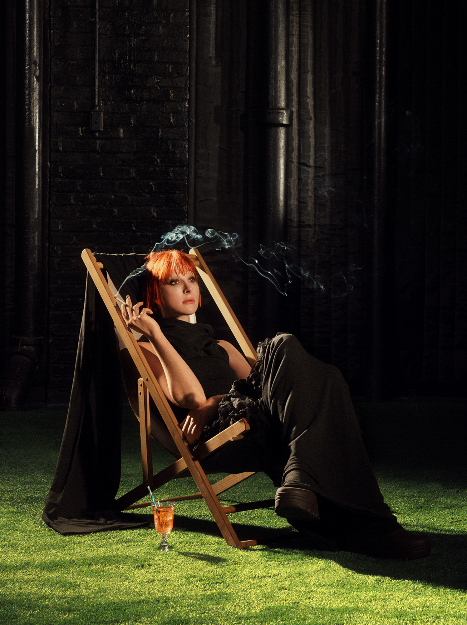 Hari Nef is seated on a lawn chair smoking a cigarette. There is an orange drink with a straw in a glass by the chair legs. The floor is covered with a green astro turf style grass. Silver appearing columns are slightly visible in the background. 