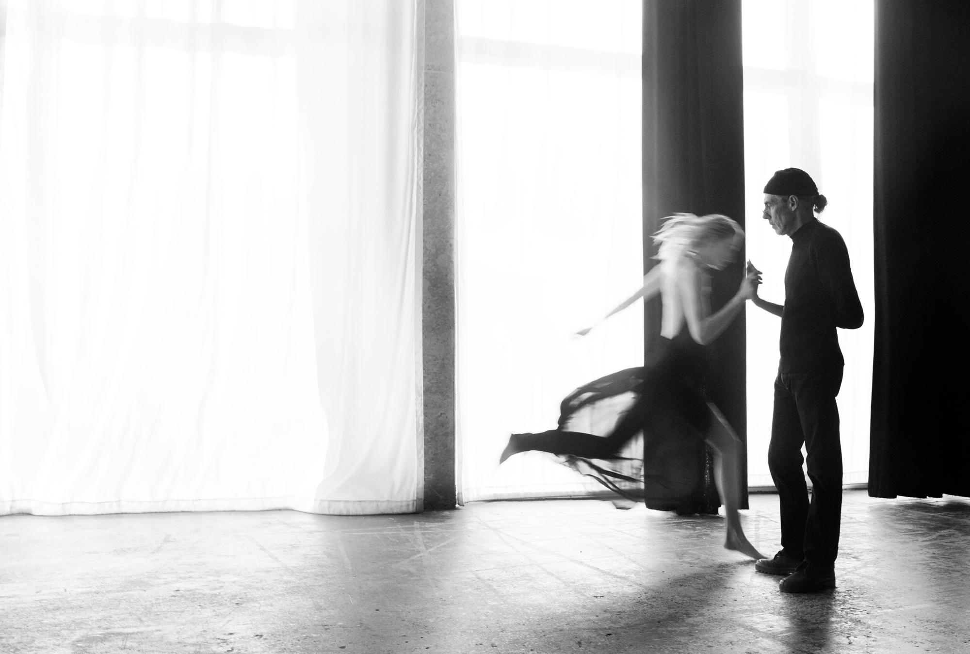 A woman is running towards a man on the right side of the frame. The woman and man are touching hands, the woman is in motion and the man is static. In the background there are sheer curtains covering the floor to ceiling windows, light is flooding the space. 