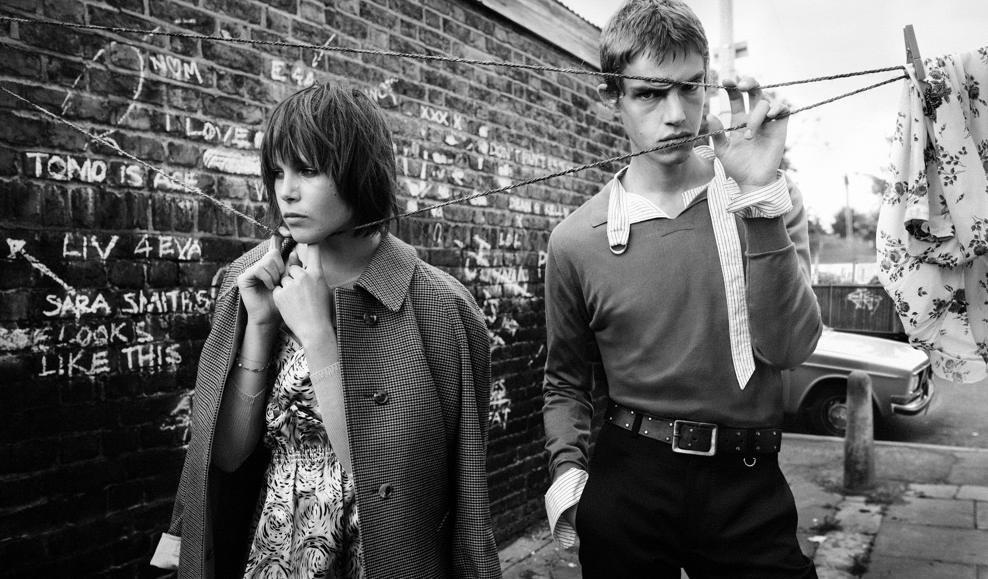 A man and a woman are standing within a clothesline in an alleyway. The man is looking at the camera while the woman is looking off to the left hand side of the frame. Chalk writings are visible on the brick wall behind them. 