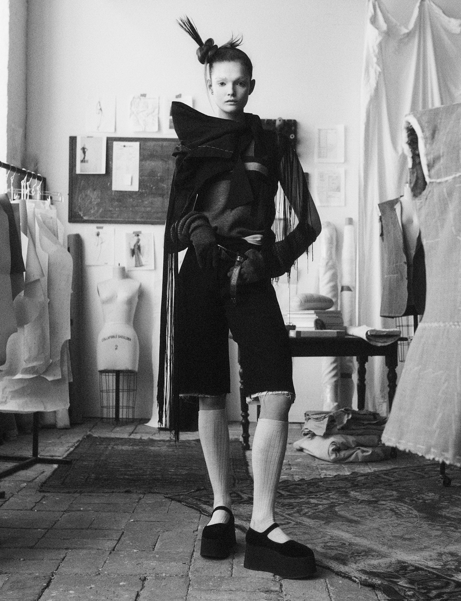 Lulu Tenney is standing in the middle of a room, which looks like an atelier. She is wearing a look that appears to be made from different sections of fabric, the edges are fraying, and there are a lot of strings. She has her eyebrows blocked out, and has her hair in a sculptural bun. 