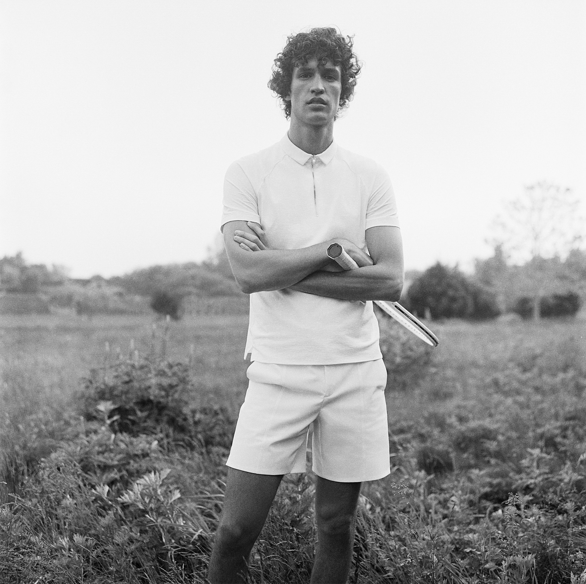 A man with curly hair is standing within some kind of field. He is wearing a short sleeve white shirt with a zipper, and white shorts. He is holding a tennis racket with his arms crossed. 