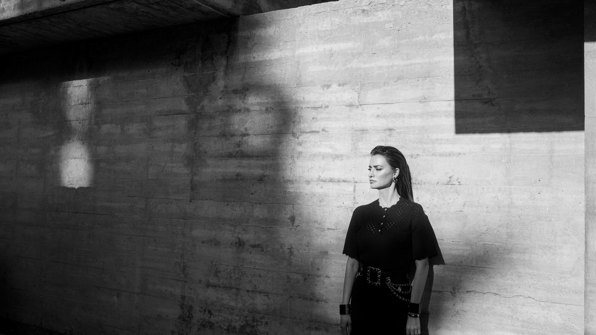 Penelope Cruz is standing in front of a concrete wall and is looking off into the distance towards the left side of the frame. She is wearing an all black look with jewelry, and a large belt buckle. 