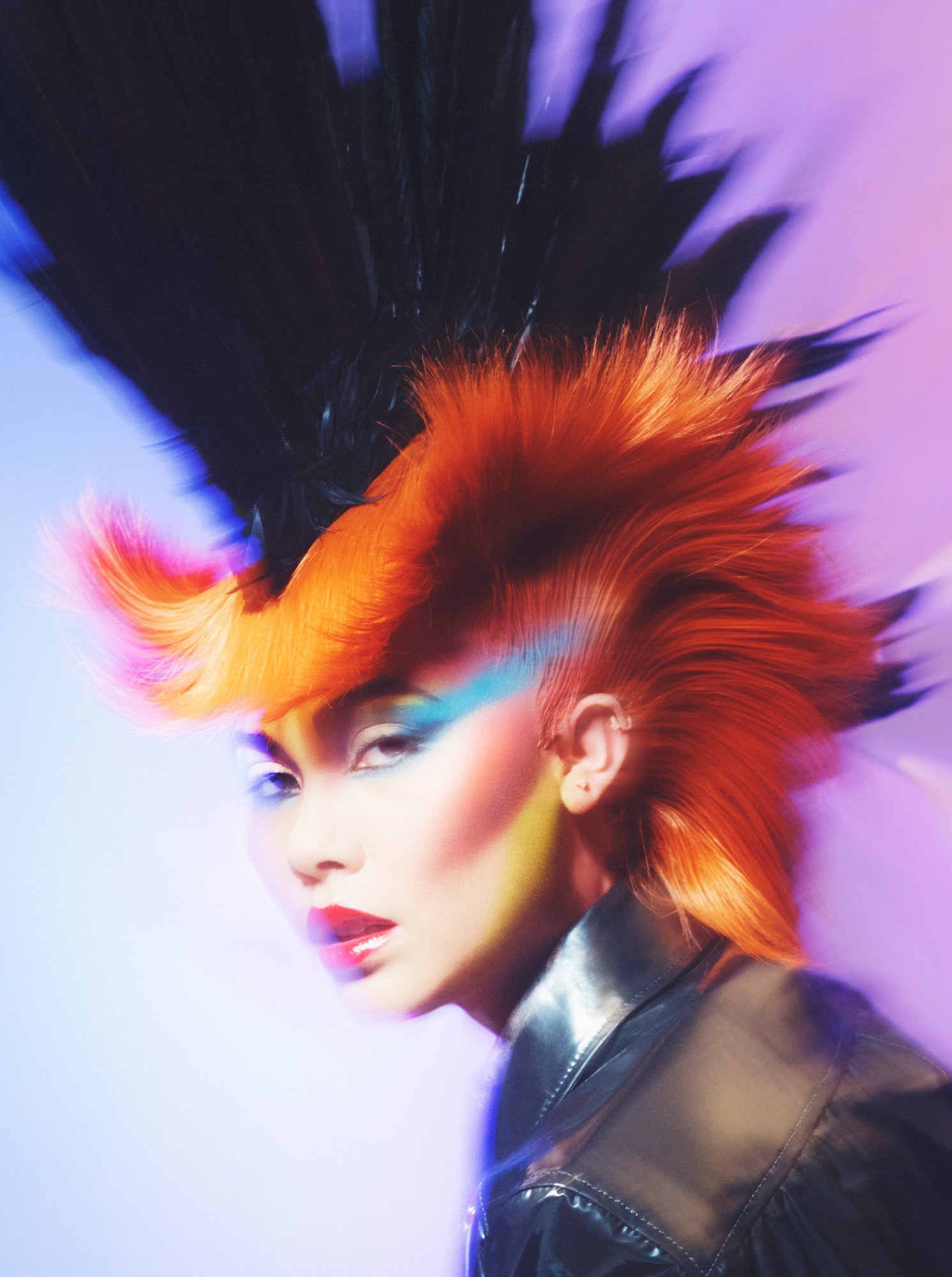 A model is seen in the image at a diagonal. The model has orange hair that is flared up at the edges, with a black looking mohawk at the top of the head. Blue angular eyeshadow, and heavy contouring are featured on the face. The model is wearing a black shiny shirt. The image has an 80s rock look and feel to it. 